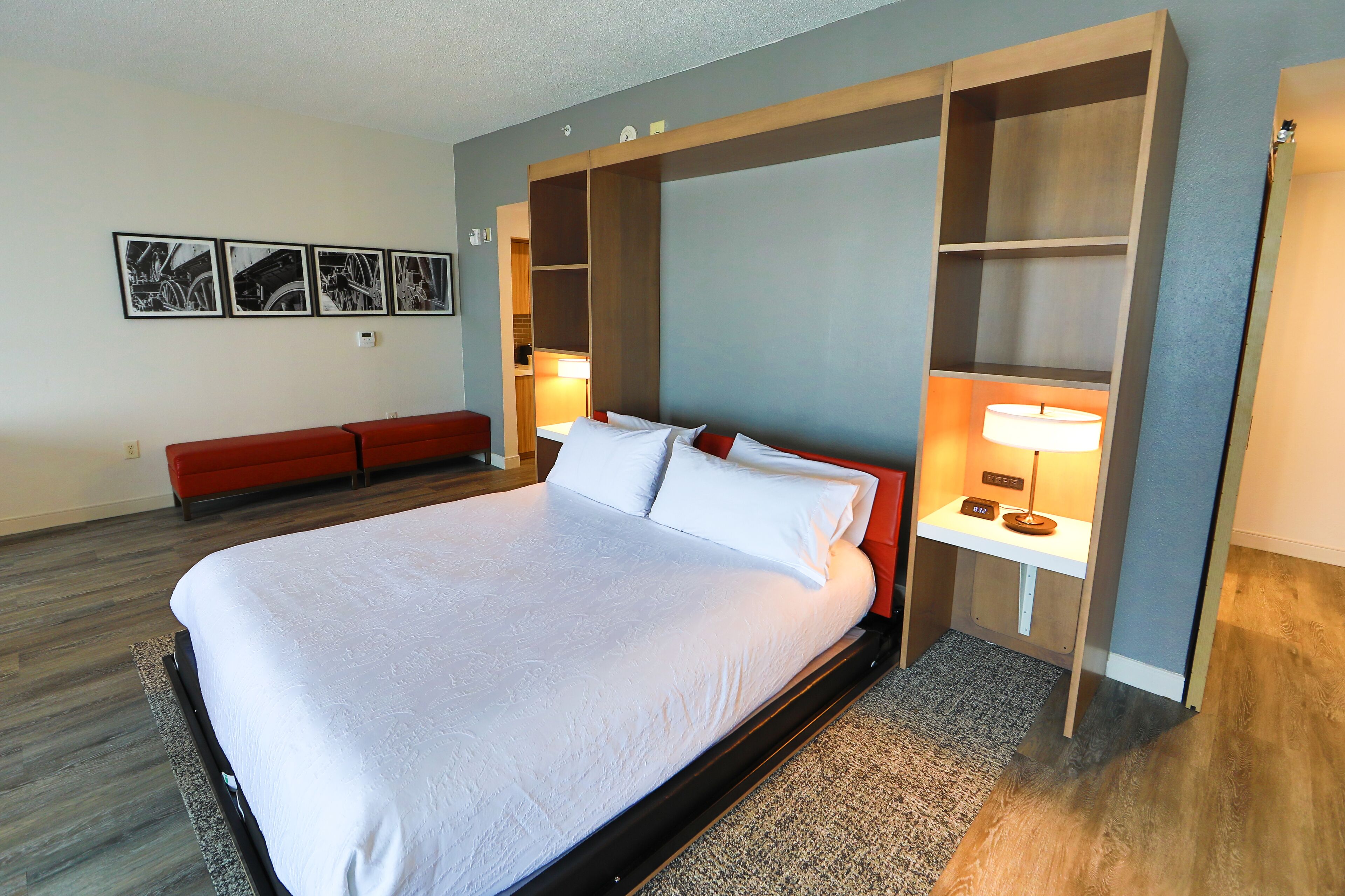Una habitación de hotel moderna con una cama grande, un banco rojo, un escritorio con una lámpara y una estantería de pared.