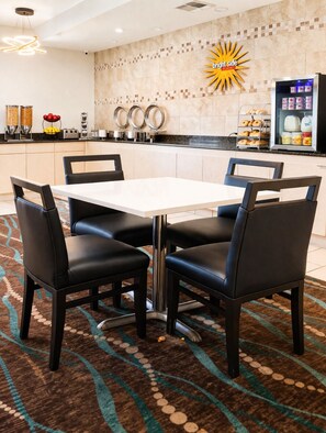 A hotel dining area with a white table and four black chairs, a patterned rug, and a breakfast display with donuts and milk.