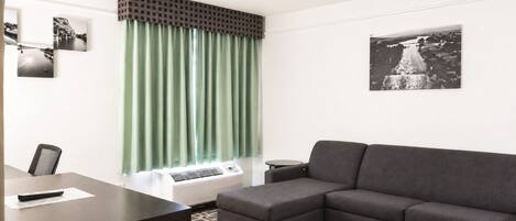 Una habitación con un sofá gris, un escritorio con una silla, una ventana con cortinas verdes y un aire acondicionado instalado en la pared.