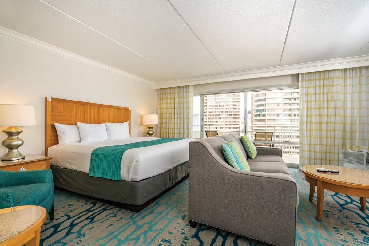 A hotel room with a large bed, a sofa, a small table, and a view of a building through the window.