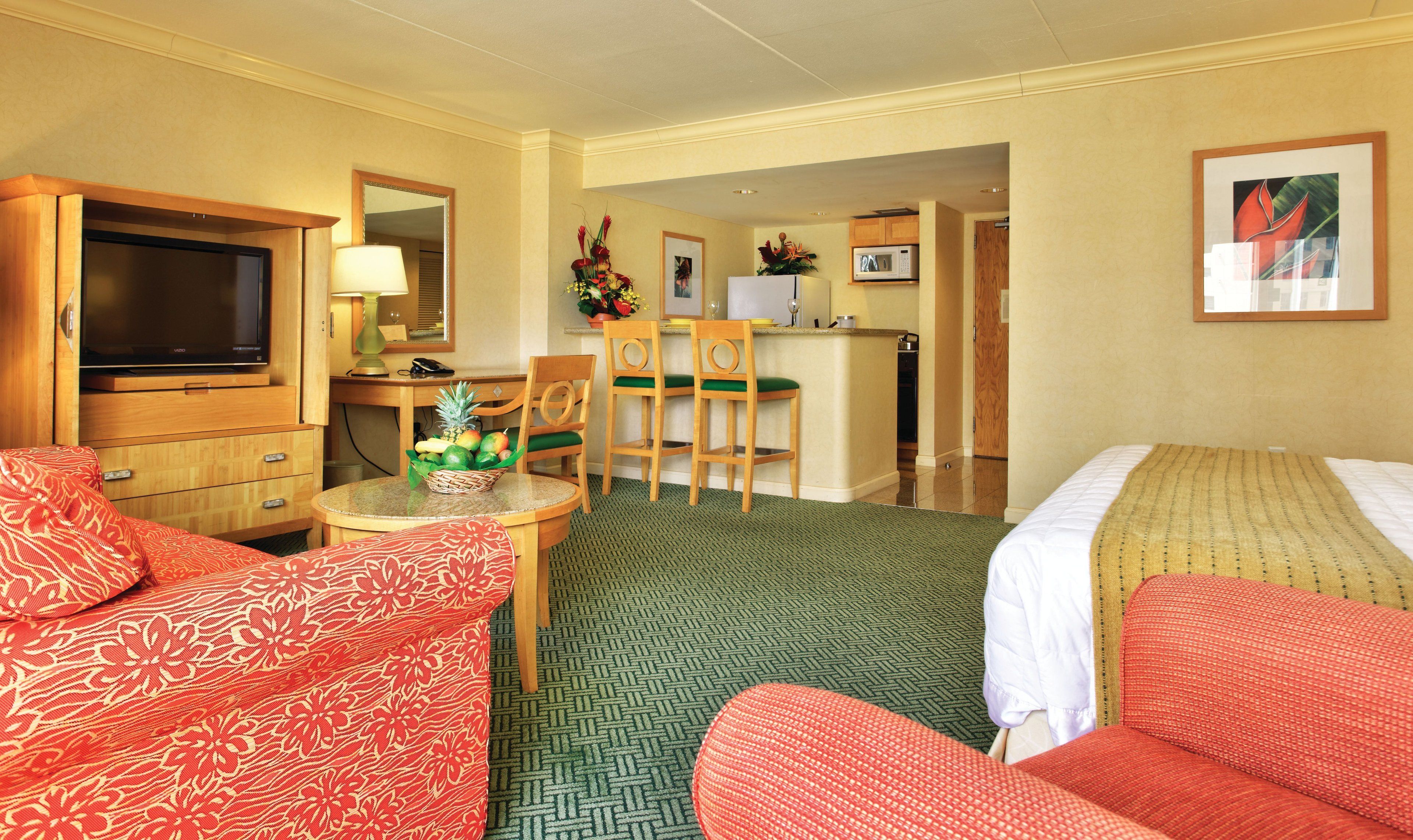 A hotel room with a television, a desk with a lamp, two chairs, a dining table with a fruit basket, and a bed with a blanket.