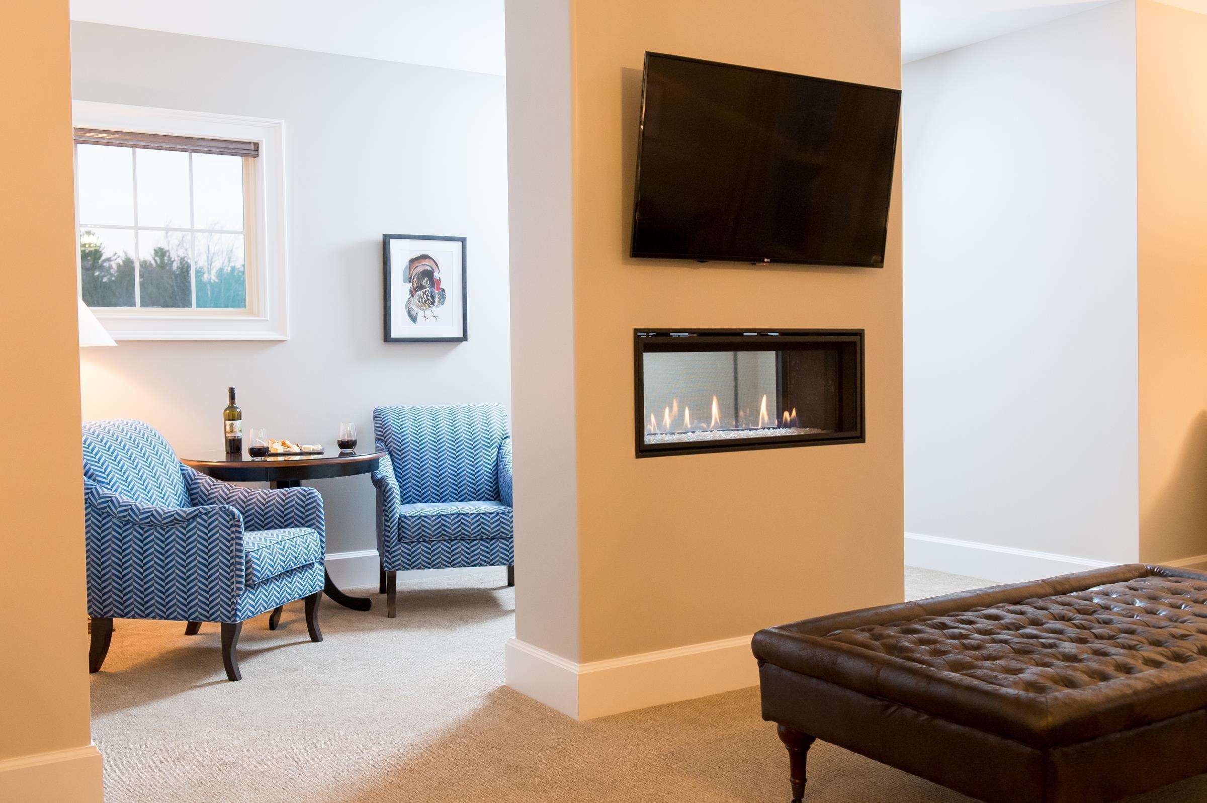 A cozy living room with a fireplace, two blue patterned armchairs, a small table with a bottle and glasses, and a television mounted on the wall.