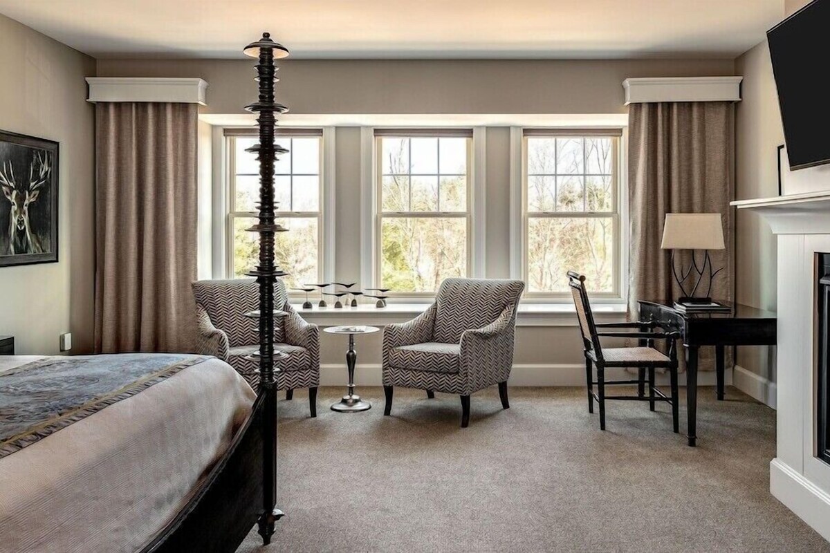 Une chambre d’hôtel avec un lit, une cheminée, un bureau avec une lampe, deux chaises, une petite table et un tableau au mur.