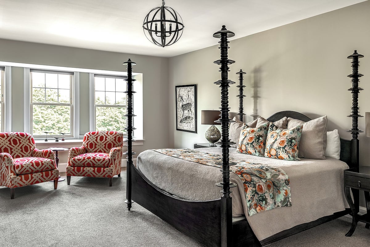 A bedroom with a four-poster bed, two armchairs, a side table, and a window with a view of trees.