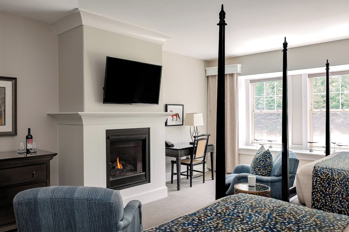 A hotel room with a fireplace, a television mounted above it, a dining area with a table and chairs, and a large window with curtains.