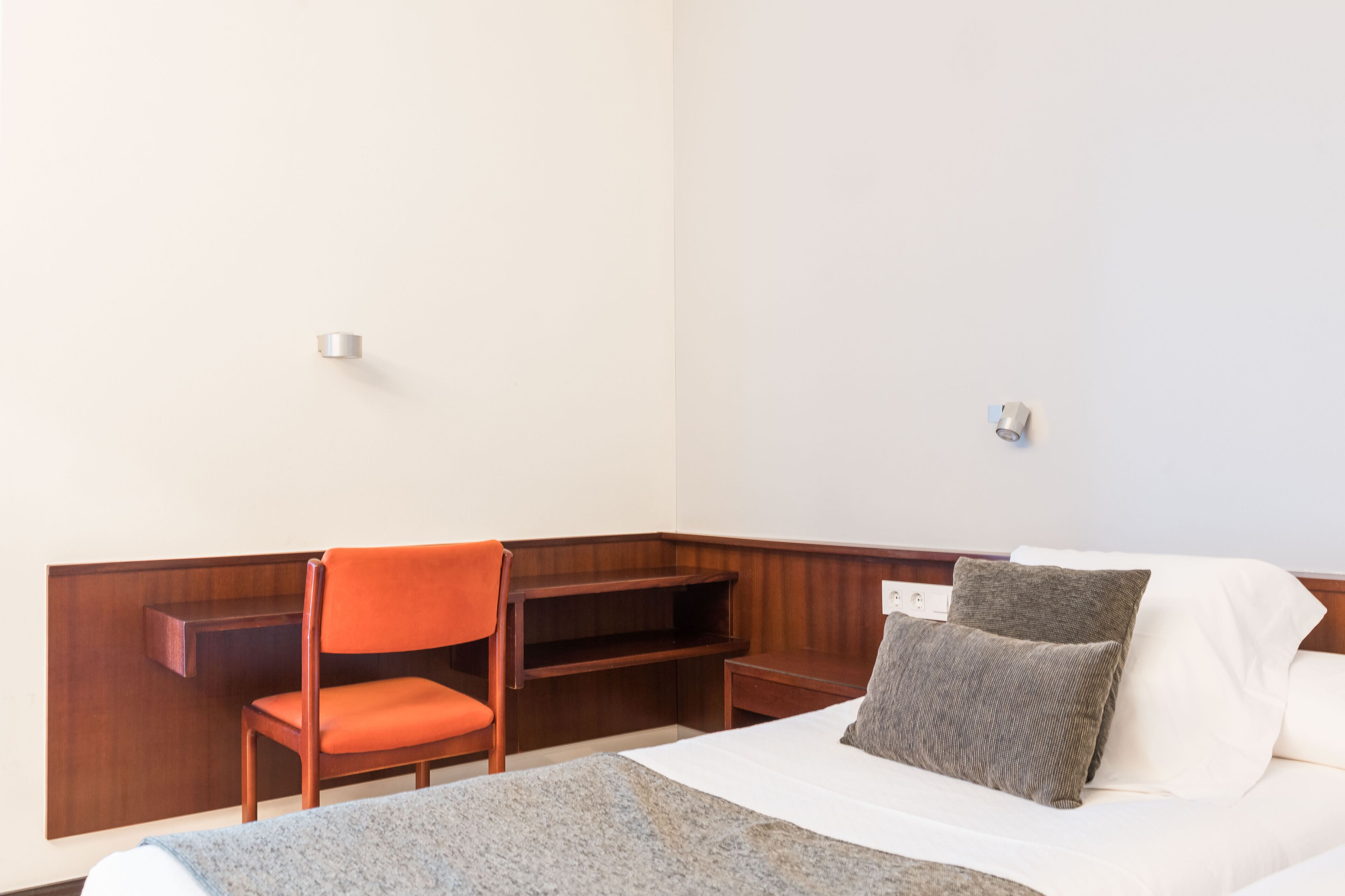 A hotel room with a bed, a wooden desk, an orange chair, and a small table.