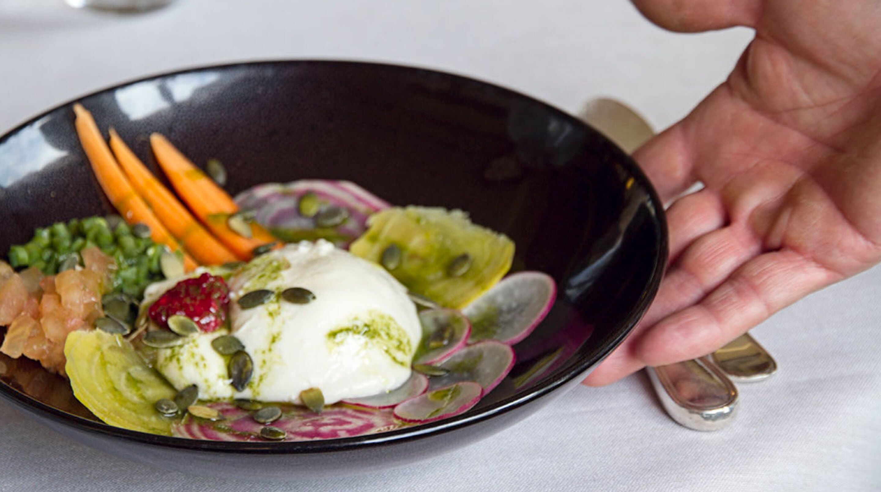 A person holding a black plate with a dessert consisting of ice cream, radishes, and pumpkin seeds.