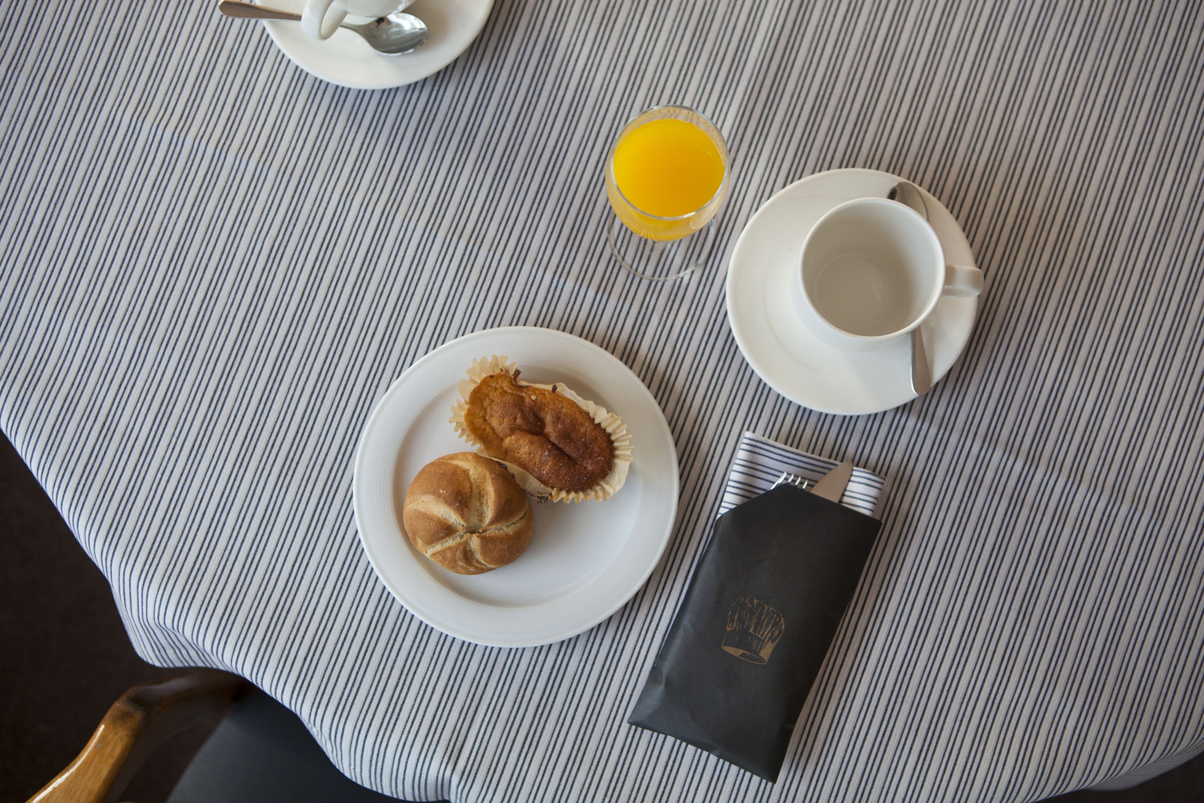 En frukostuppdukning med en randig duk, en tallrik med två bakverk, ett glas apelsinjuice, en kopp med tillhörande fat och en servett med en logotyp.