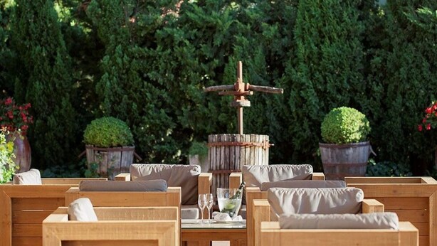 A wooden outdoor seating area with cushioned benches and a central wooden structure with a bottle and glasses. - Hotel Empordà (Figueres)