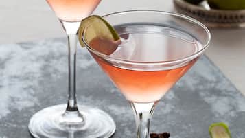 Two martini glasses with a pinkish drink and lime garnish on a marble surface.