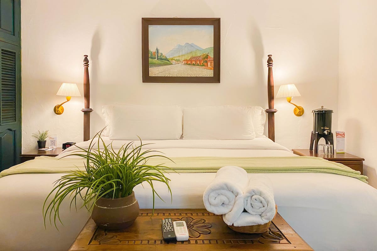 Una cama bien tendida con sábanas blancas, una planta en maceta, un control remoto y una cesta de toallas sobre una mesita de noche de madera.