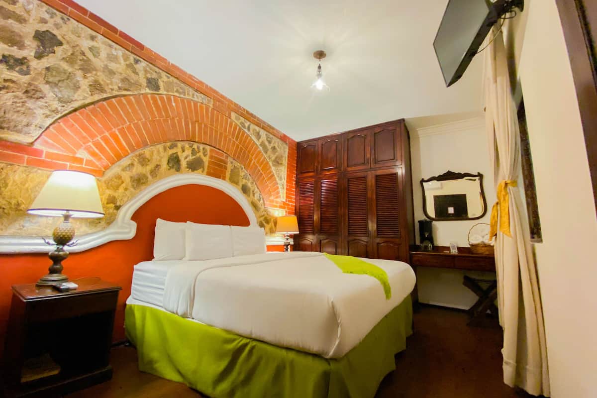 Una habitación de hotel con un arco de ladrillo, una cama con ropa de cama verde y blanca, una mesita de noche con una lámpara, un armario de madera, un espejo y un televisor montado en la pared.