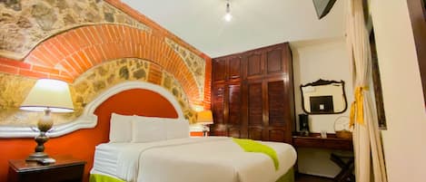 A hotel room with a brick arch, a bed with green and white bedding, a nightstand with a lamp, a wooden wardrobe, a mirror, and a TV mounted on the wall.