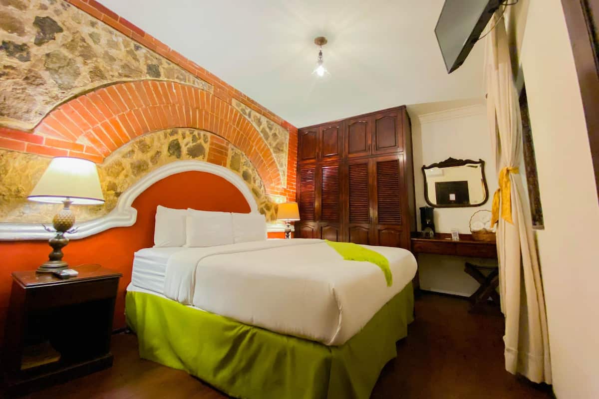 Hotelkamer met een bakstenen boog, een bed met groen-witte beddengoed, een nachtkastje met een lamp, een houten garderobe, een spiegel en een aan de muur bevestigde televisie.