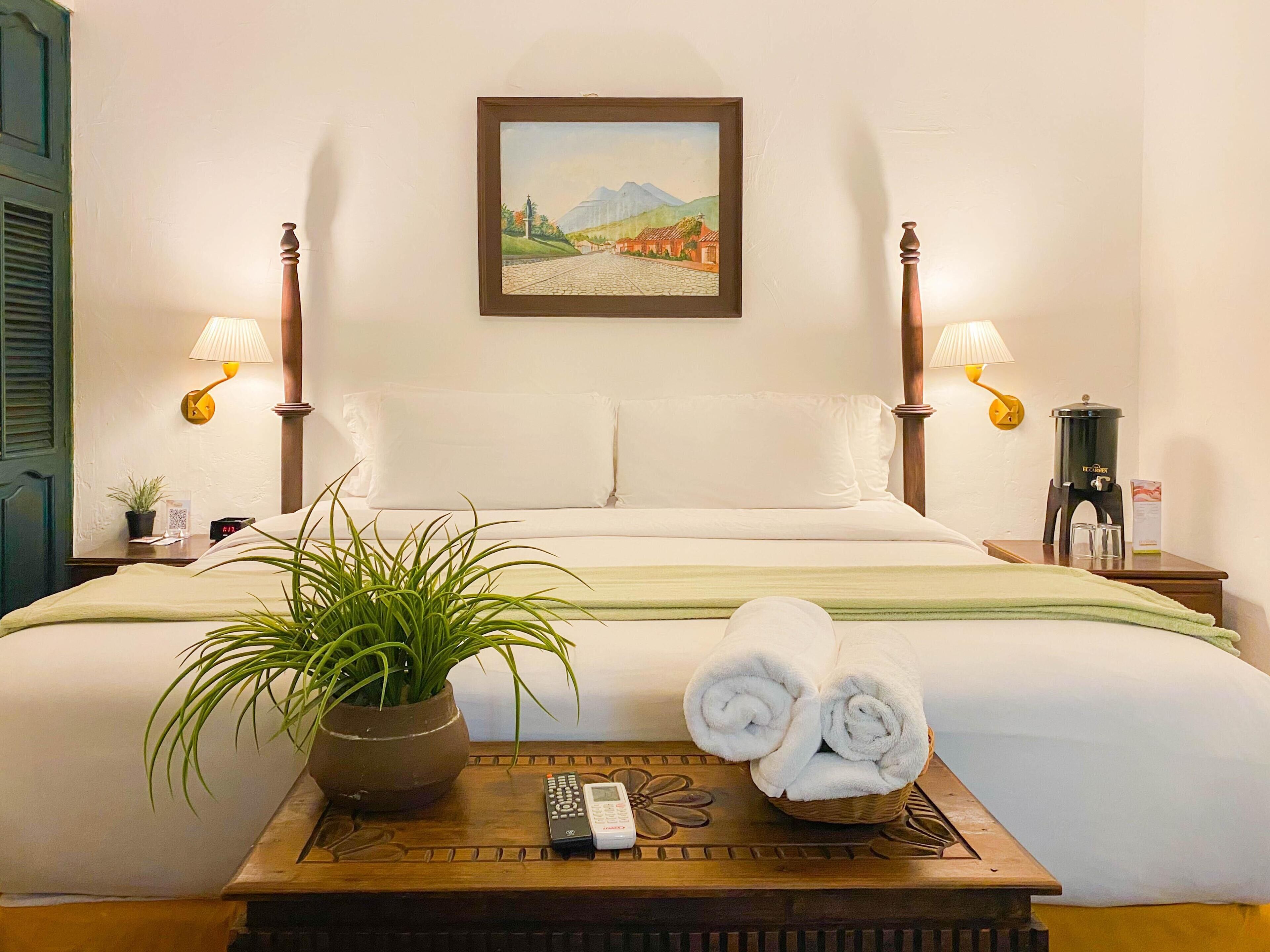 A hotel room with a bed, two bedside lamps, a painting, a potted plant, a tray with towels and a remote control, and a coffee maker.