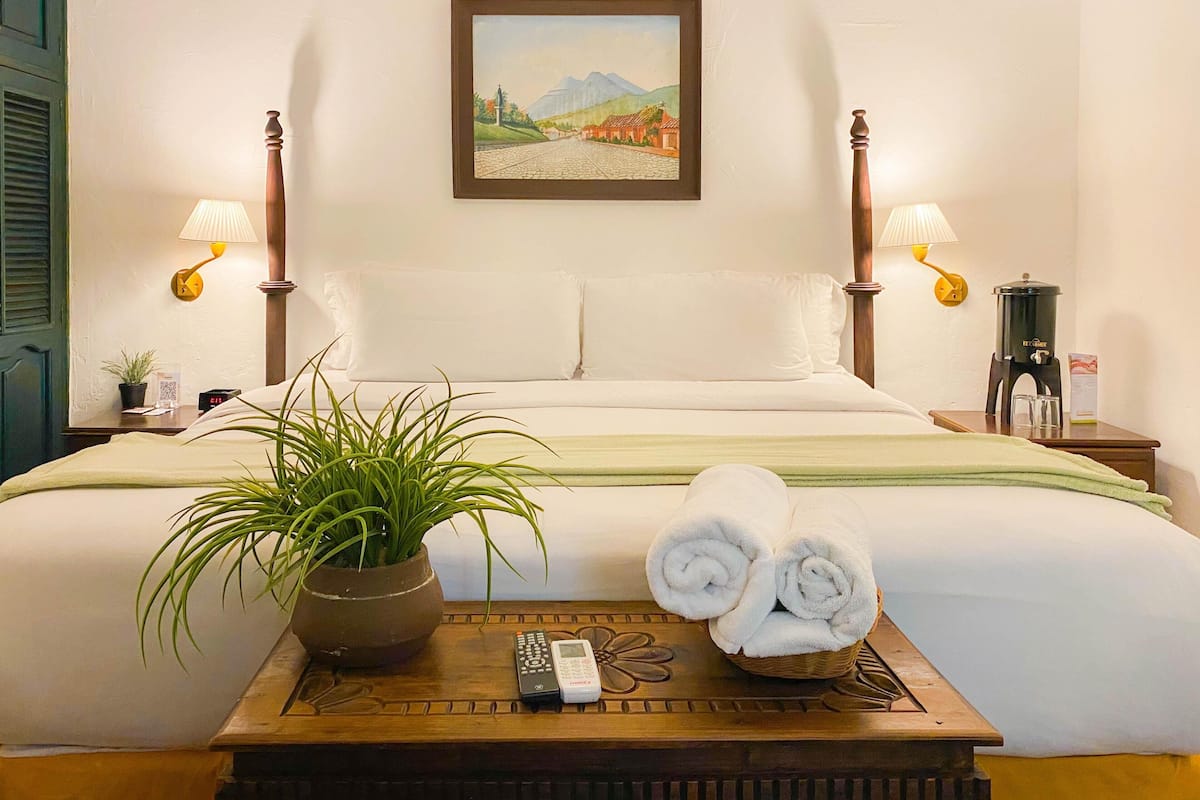 Habitación de hotel con cama, dos lámparas de noche, un cuadro, una planta en maceta, una bandeja con toallas y un control remoto, y una cafetera.