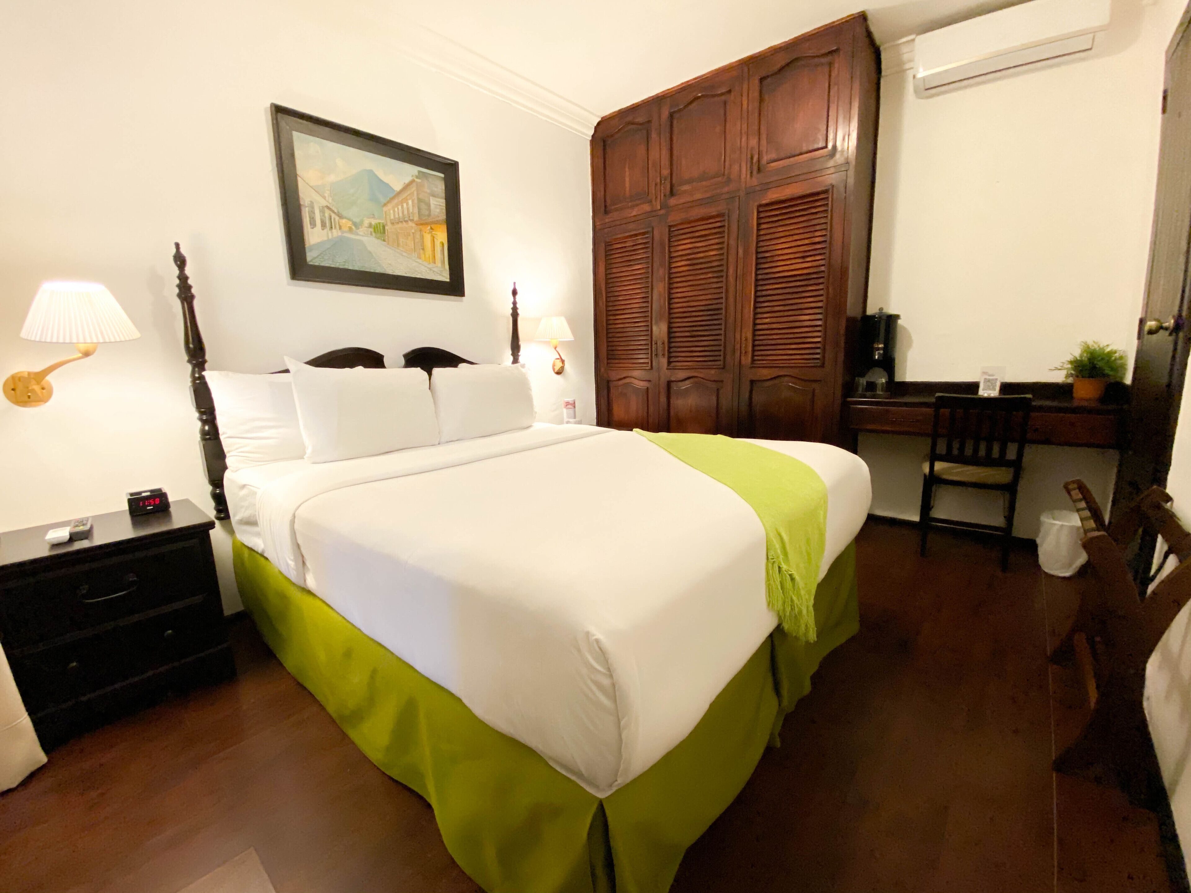 A neatly made bed with a green bed skirt, a wooden wardrobe, a desk with a chair, and a painting on the wall.