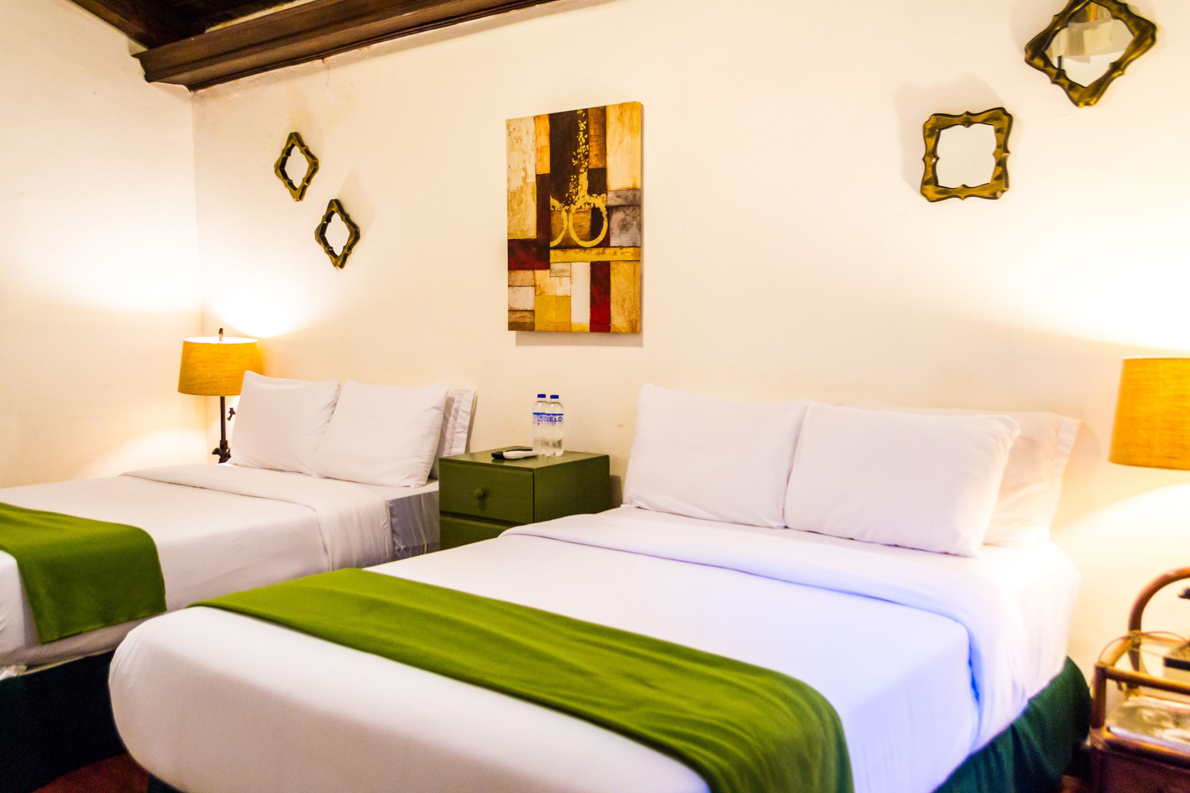 Habitación de hotel con dos camas, cada una con ropa blanca y almohadones verdes. Hay un cuadro en la pared y una mesita con una lámpara.