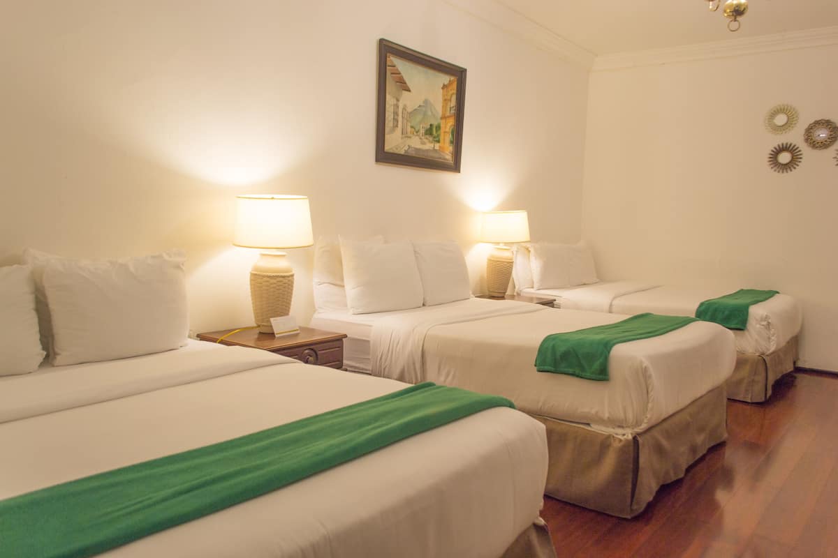 Een hotelkamer met drie bedden, elk voorzien van wit beddengoed en groene plaids. Twee nachtkastjes met lampen staan er, en aan de muur hangt een schilderij.