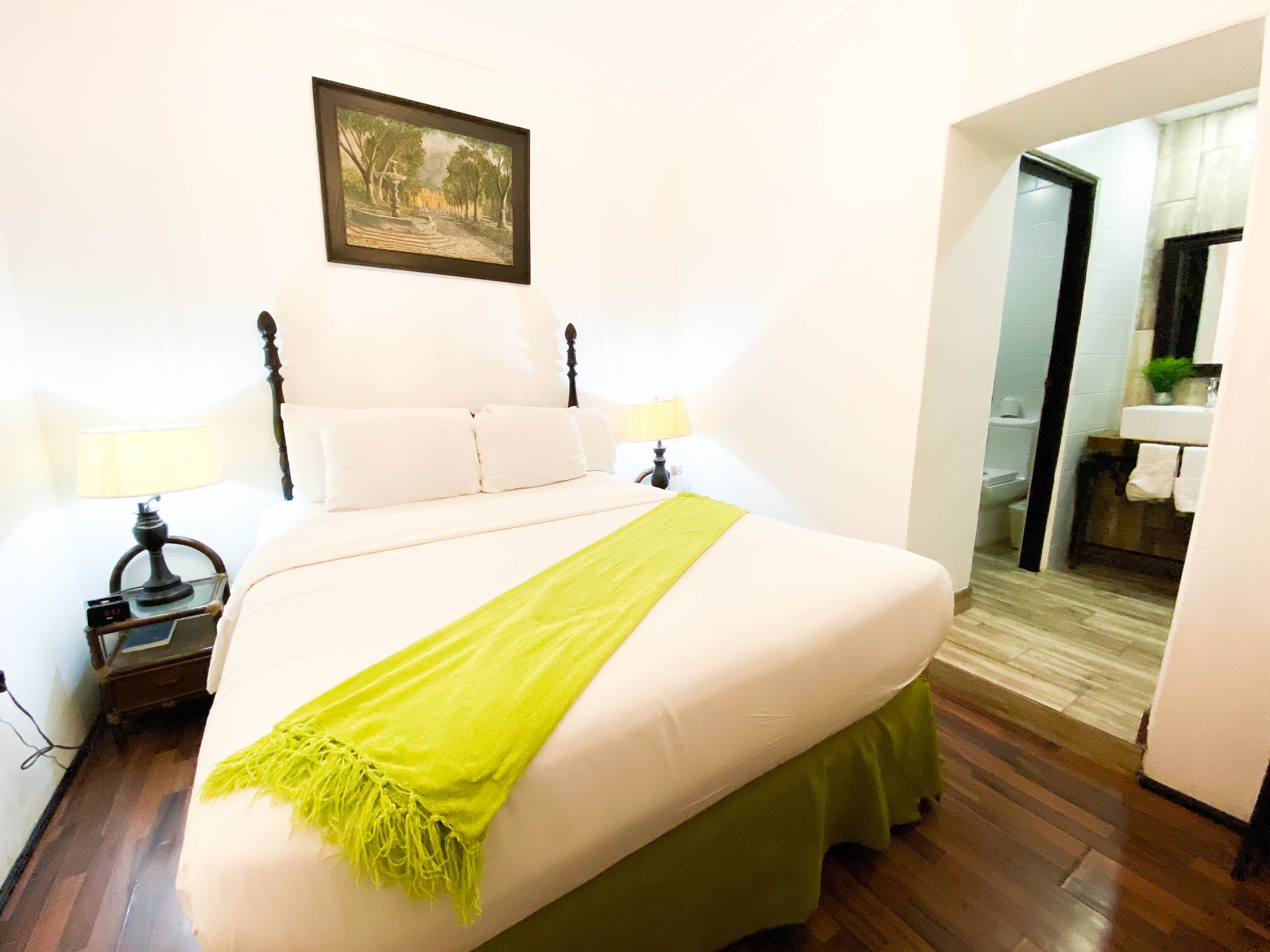 Una cama bien hecha con un edredón verde, dos lámparas de noche y un cuadro en la pared.