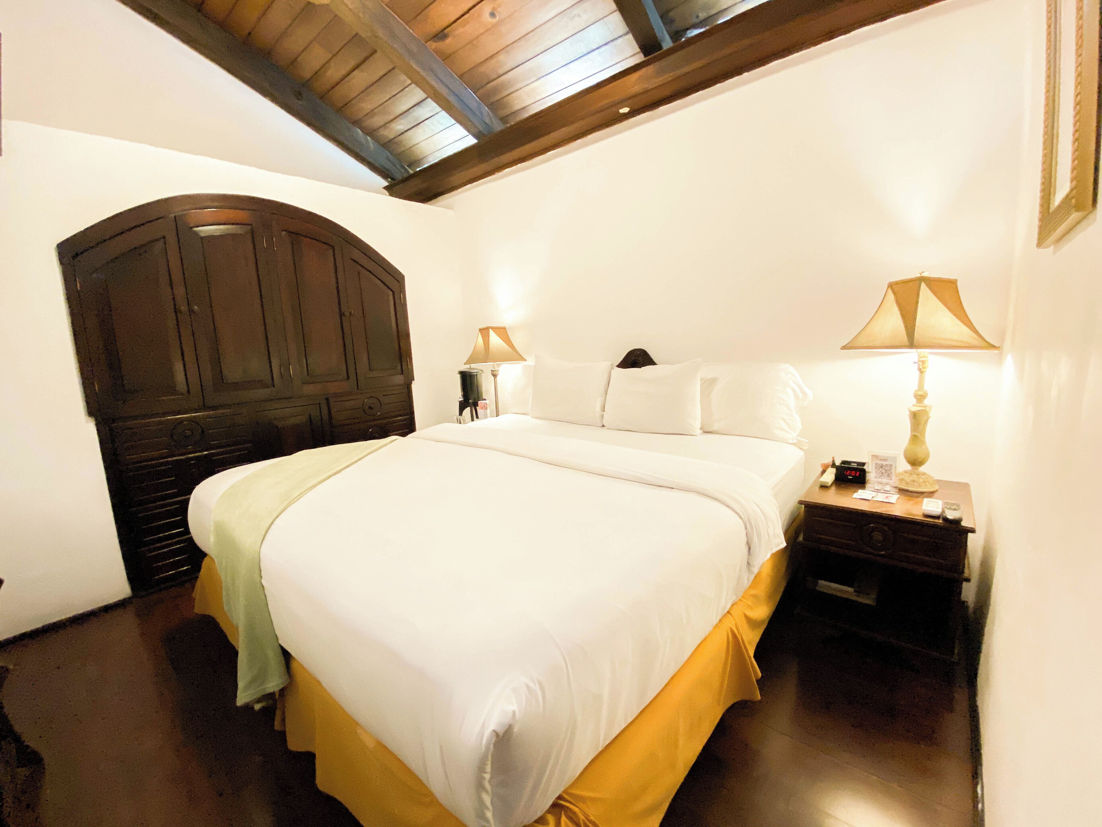 Un dormitorio con una cama grande, cabecera de madera, dos mesitas de noche con lámparas y techo de madera.