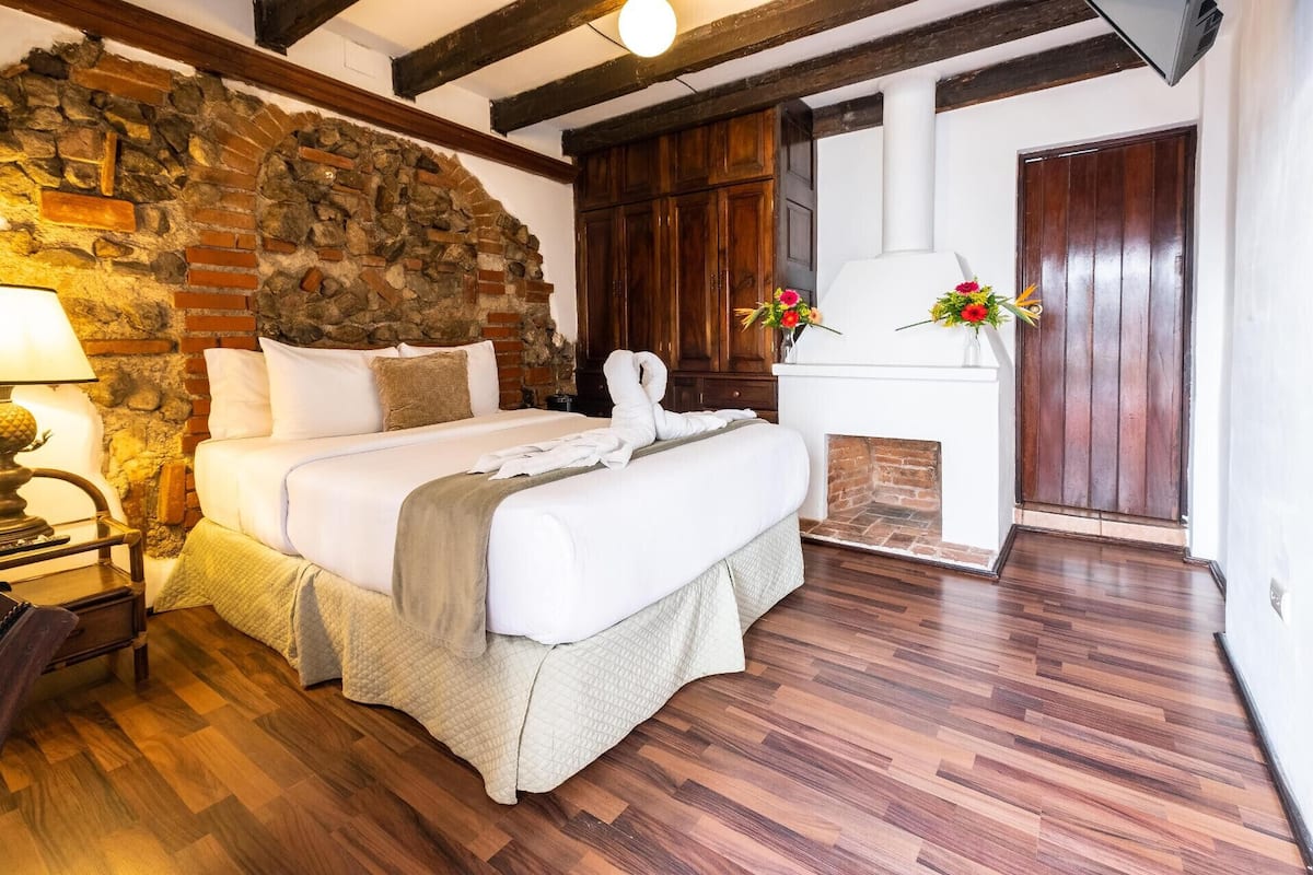 Un dormitorio con pared de piedra, vigas de madera, una cama con sábanas blancas, una mesita de noche, una lámpara y una chimenea decorada con flores.