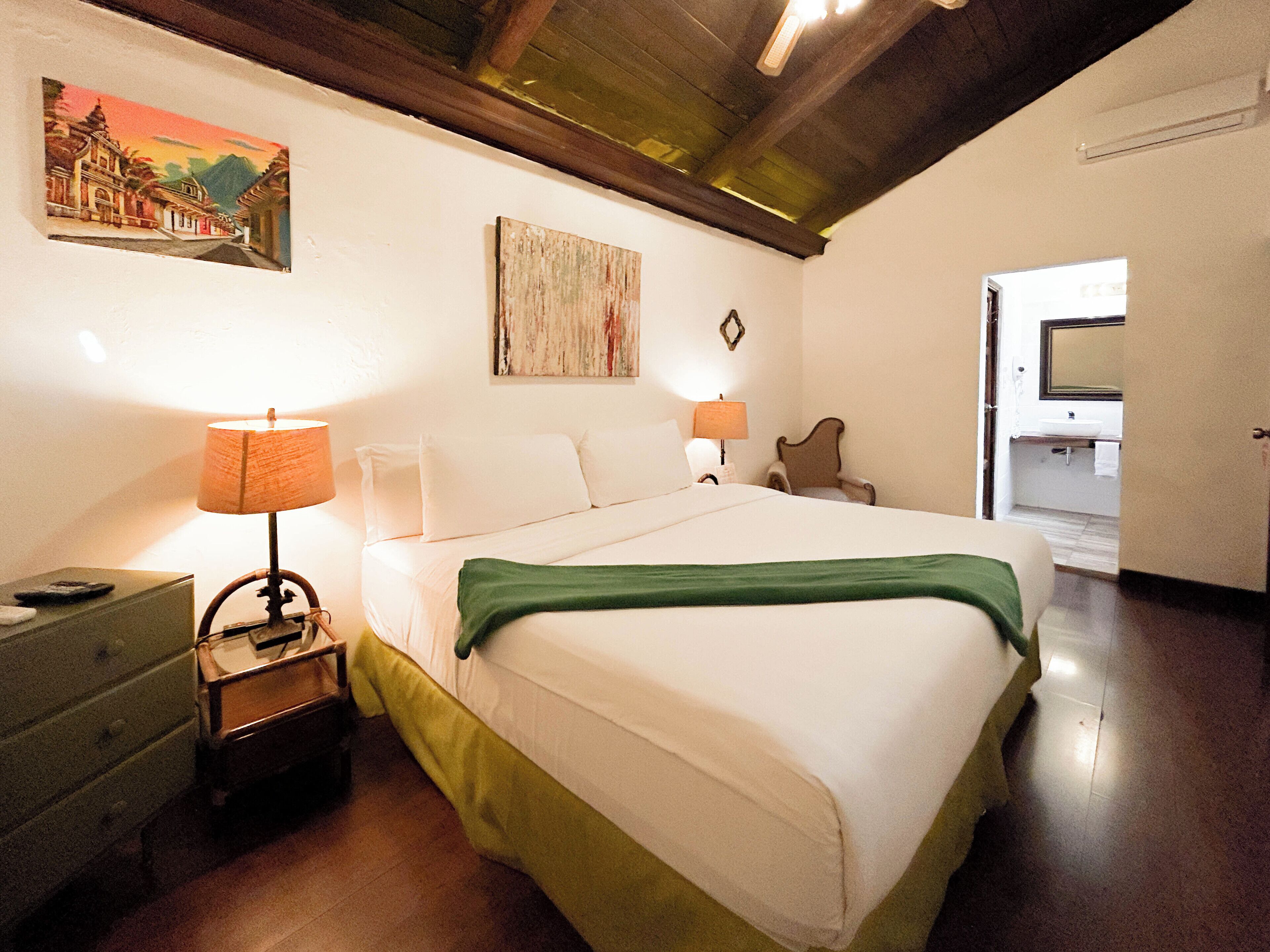 A hotel room with a bed, two bedside lamps, a nightstand, a chair, a painting on the wall, and a bathroom visible through an open door.