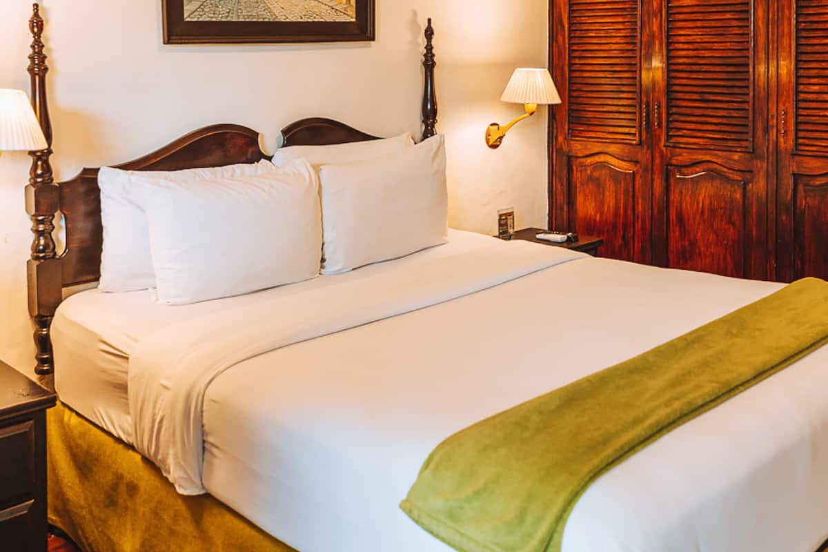 Una cama bien hecha con sábanas blancas y una manta verde, cabecera de madera, dos lámparas de noche y un armario de madera con puertas empañadas.