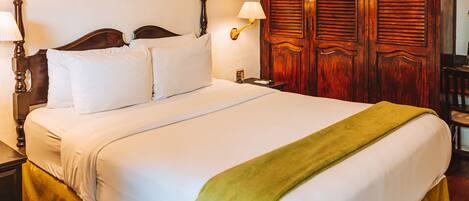 A neatly made bed with white linens and a green throw blanket, wooden headboard, two bedside lamps, and a wooden wardrobe with louvered doors.