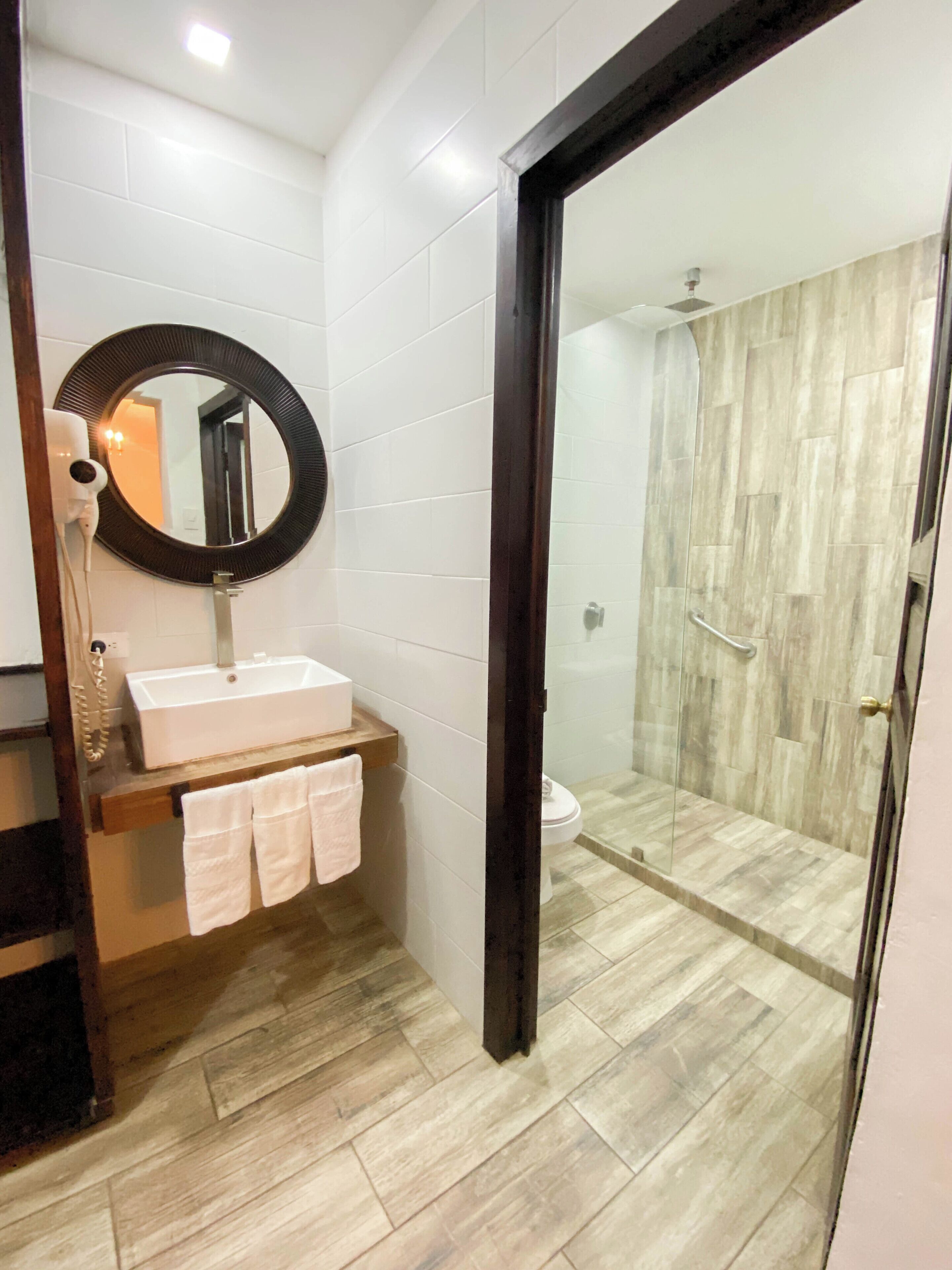 Un baño moderno con un lavamanos blanco, un espejo redondo y una zona de ducha con paredes de vidrio.