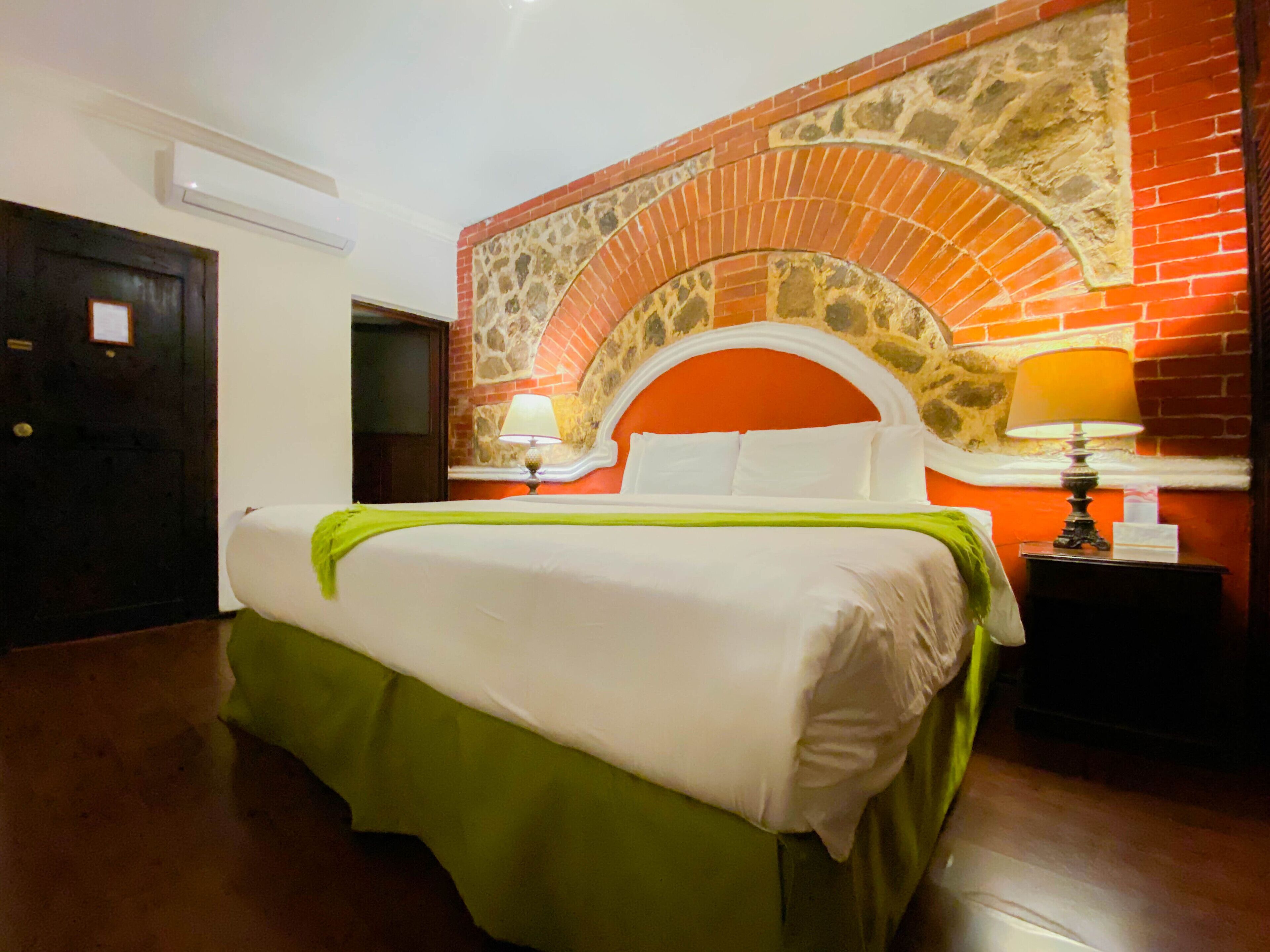 A hotel room with a brick fireplace, a bed with green and white bedding, and two bedside lamps.