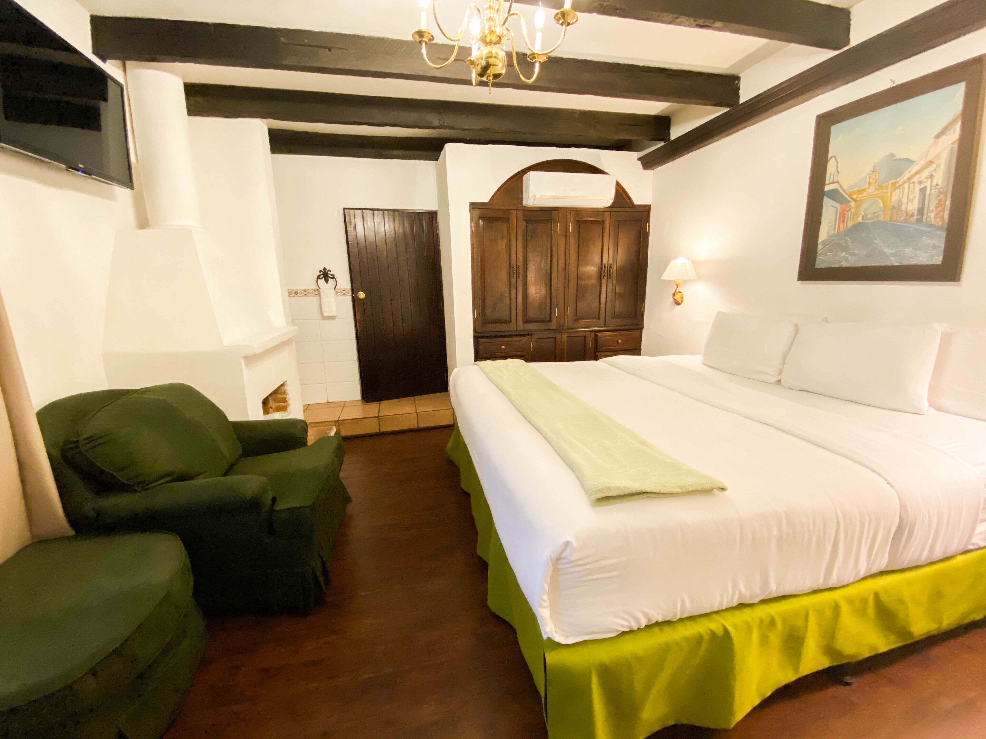 A hotel room with a bed, two green armchairs, a wooden wardrobe, a painting on the wall, and a chandelier.