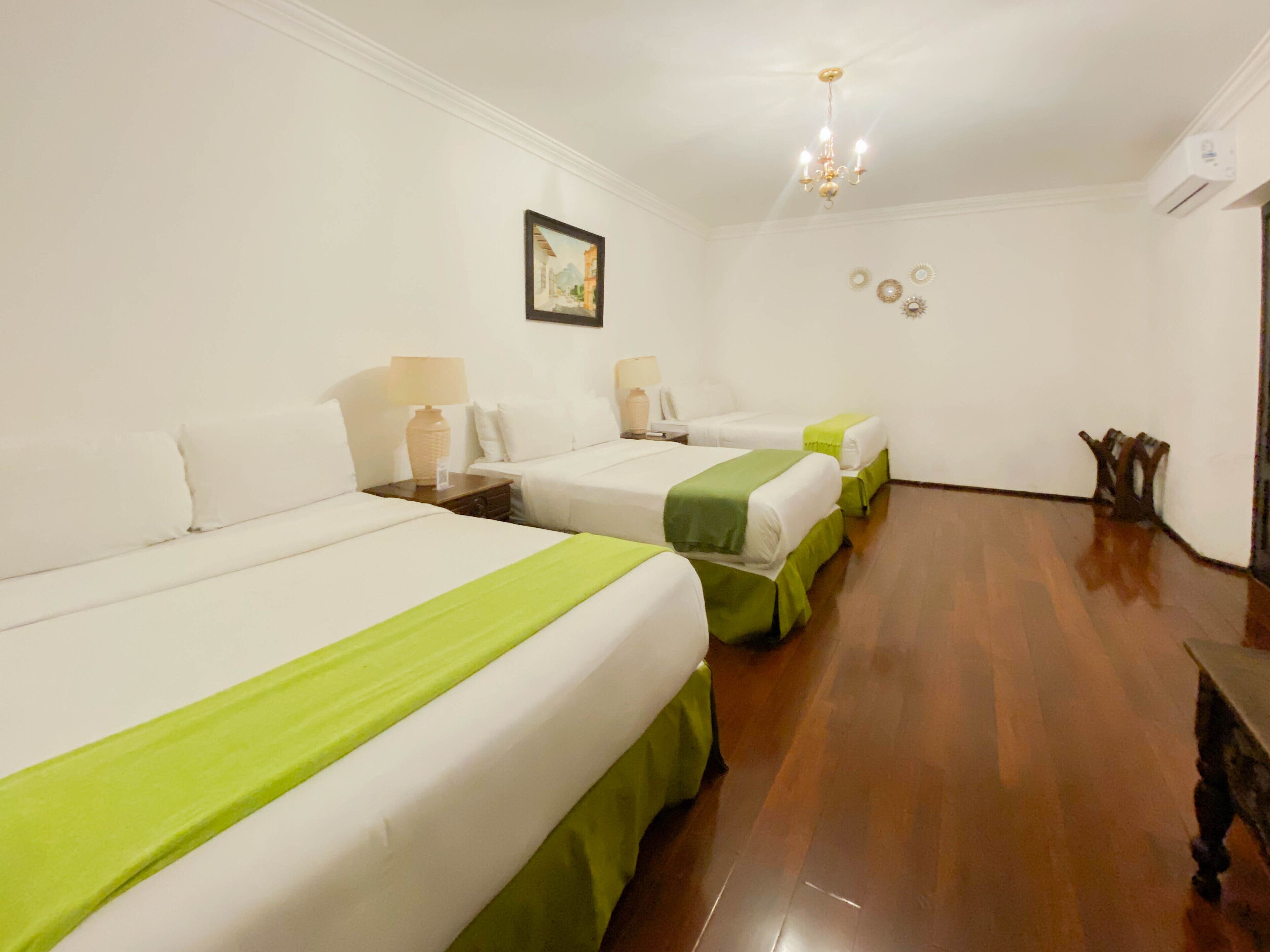 Une chambre d’hôtel avec trois lits simples, chacun recouvert d’une couvre-lit verte, avec du linge de lit blanc et des tables de chevet dotées de lampes. Un lustre orne le plafond, un tableau encadré est accroché au mur, et un banc en bois se trouve près de la porte.