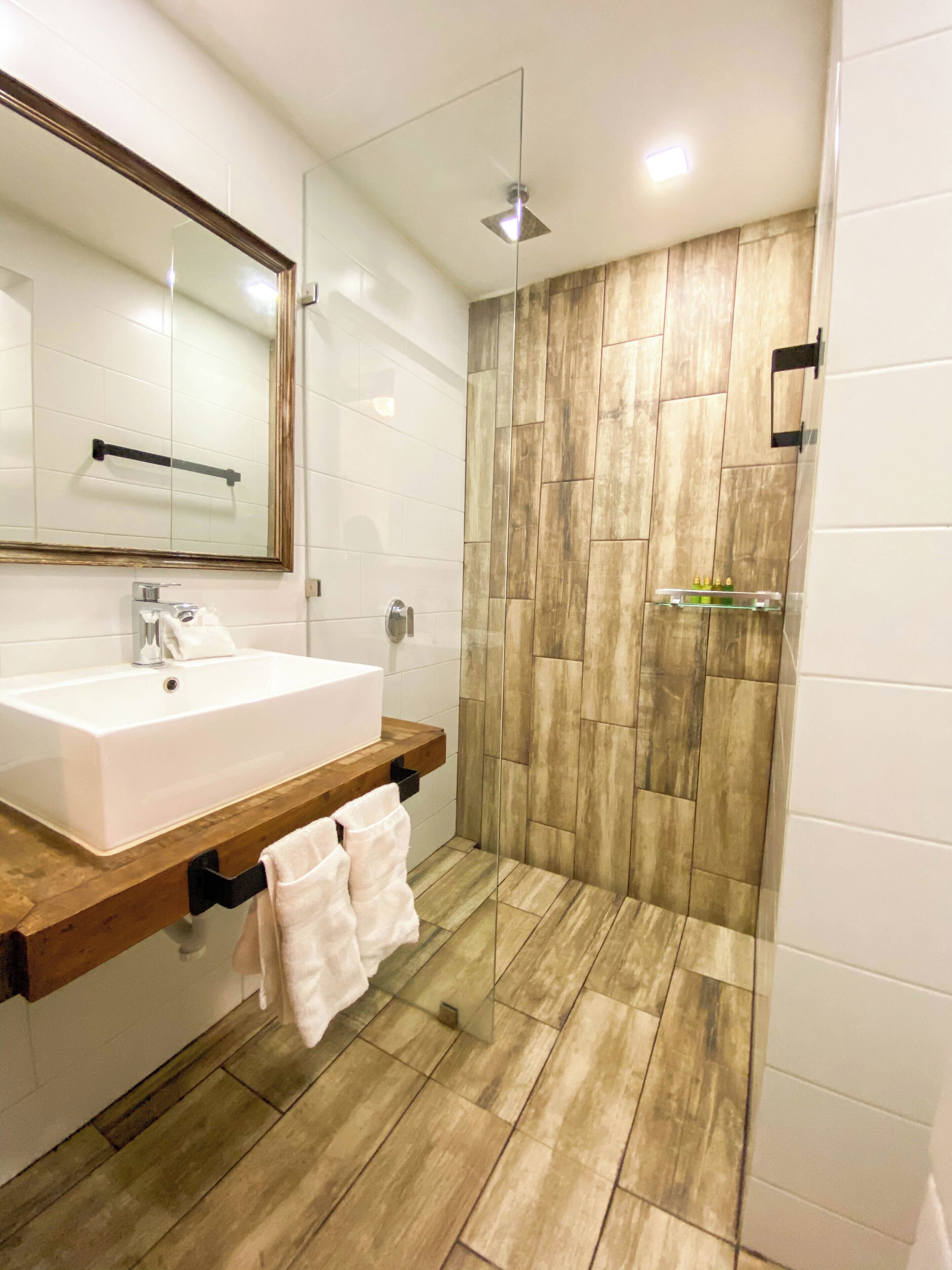 A modern bathroom with a large mirror, white sink, wooden vanity, and a shower area with wooden tiles.