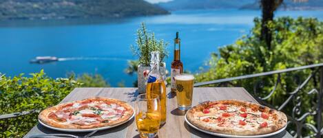 A table with two pizzas, a beer glass, and a bottle of beer, overlooking a lake with a boat in the distance.