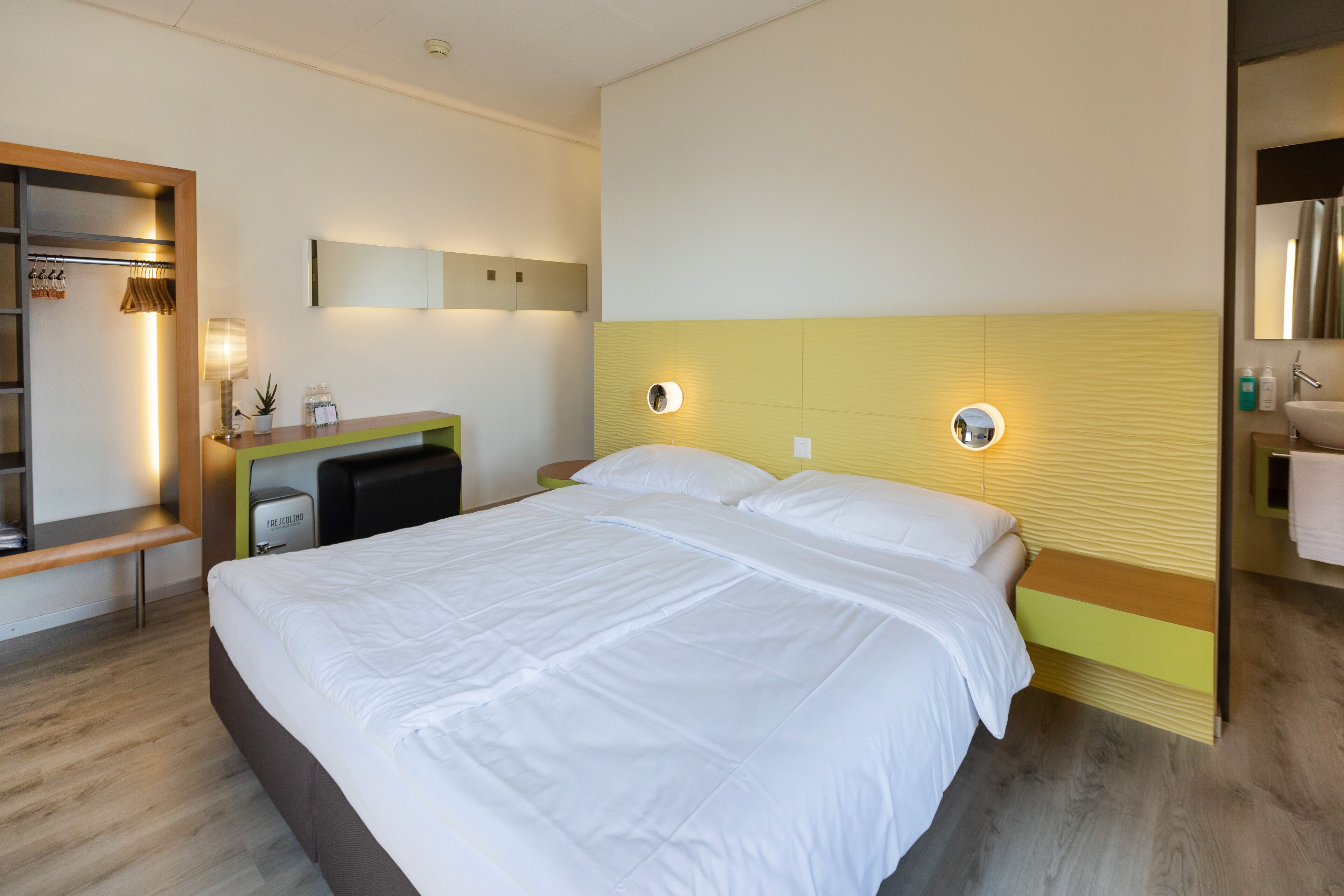 A hotel room with a large bed, a desk with a lamp, a mirror, and a bathroom with a sink and towel rack.