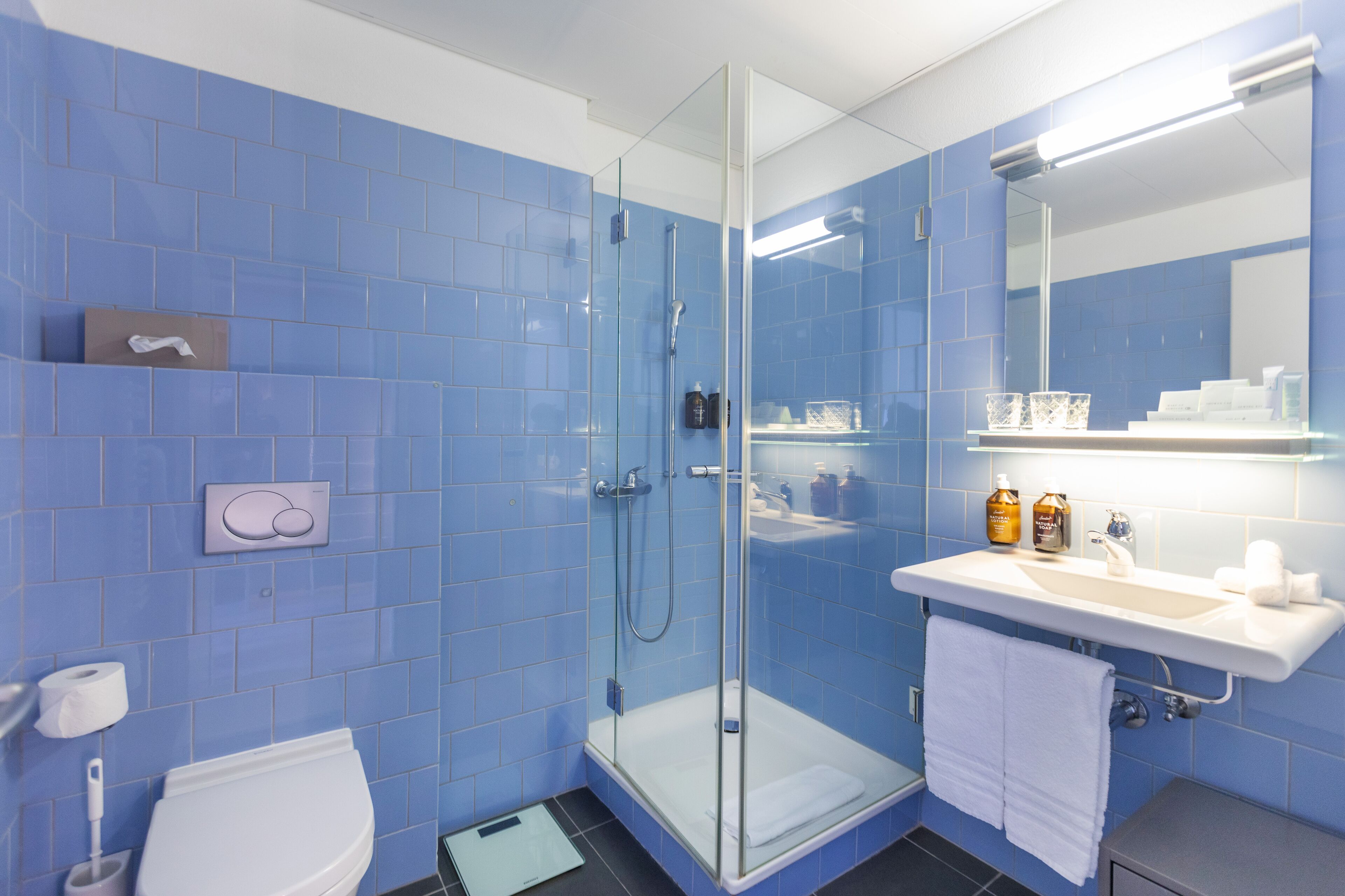 Un baño con azulejos azules, cabina de ducha con vidrio, lavamanos blanco con artículos de aseo, y un inodoro.