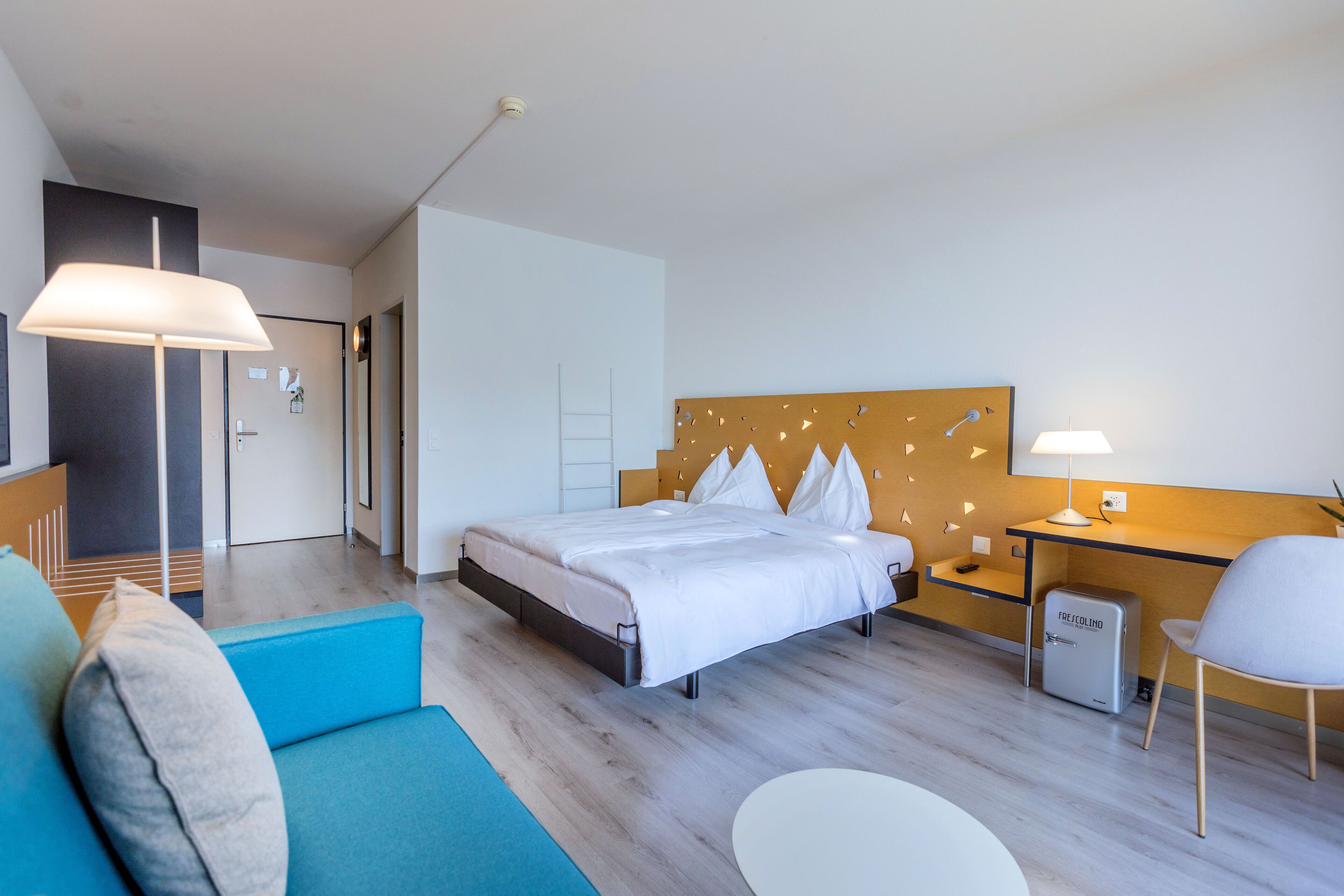 Une chambre d’hôtel moderne équipée d’un lit, d’un bureau, d’une chaise, d’une lampe et d’un petit réfrigérateur.