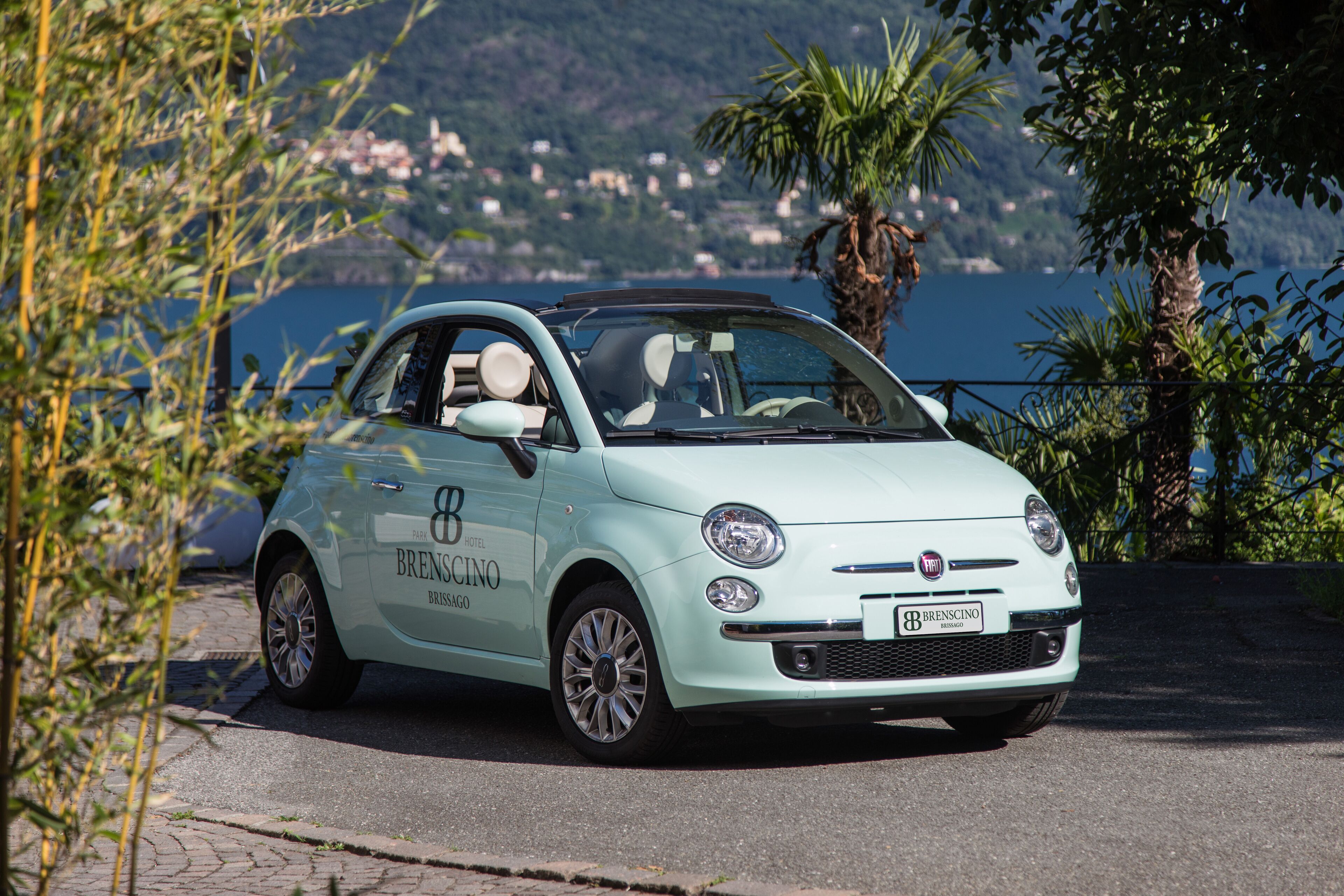 A light blue car with "BRESCINO" branding parked on a paved area with a scenic coastal background.