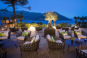 An outdoor seating area with wicker furniture and a central tree, overlooking a body of water and mountains. - Parkhotel Brenscino Brissago (Brissago)