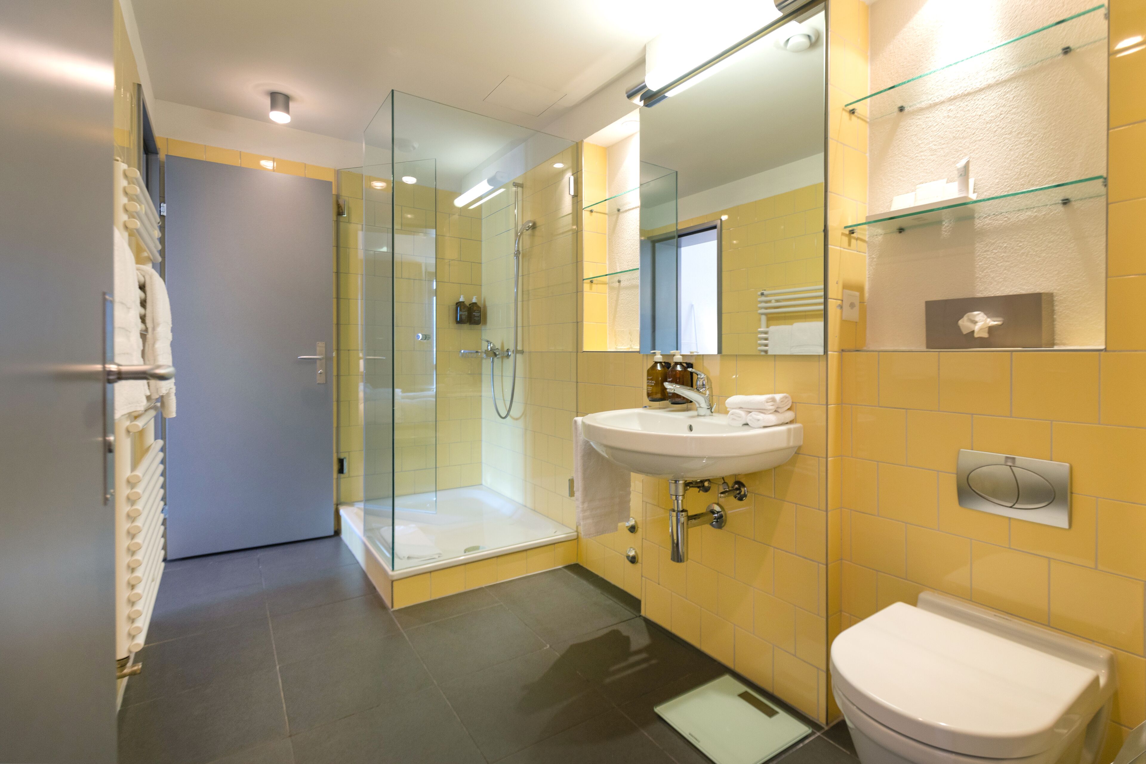 Een moderne badkamer met een douche in een glazen cabine, een witte wastafel en een gele betegelde muur.