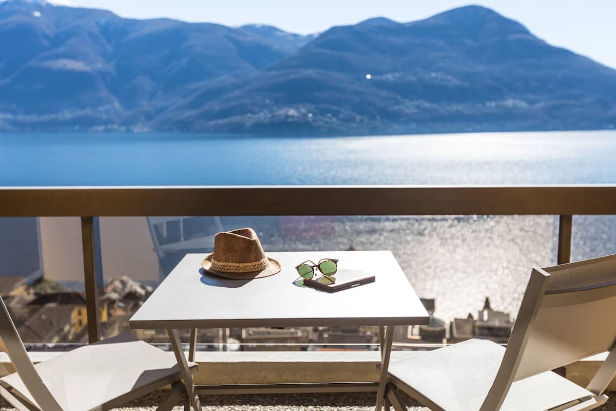 Un balcón con una mesa, un sombrero y gafas de sol, con vistas a un lago tranquilo y montañas.