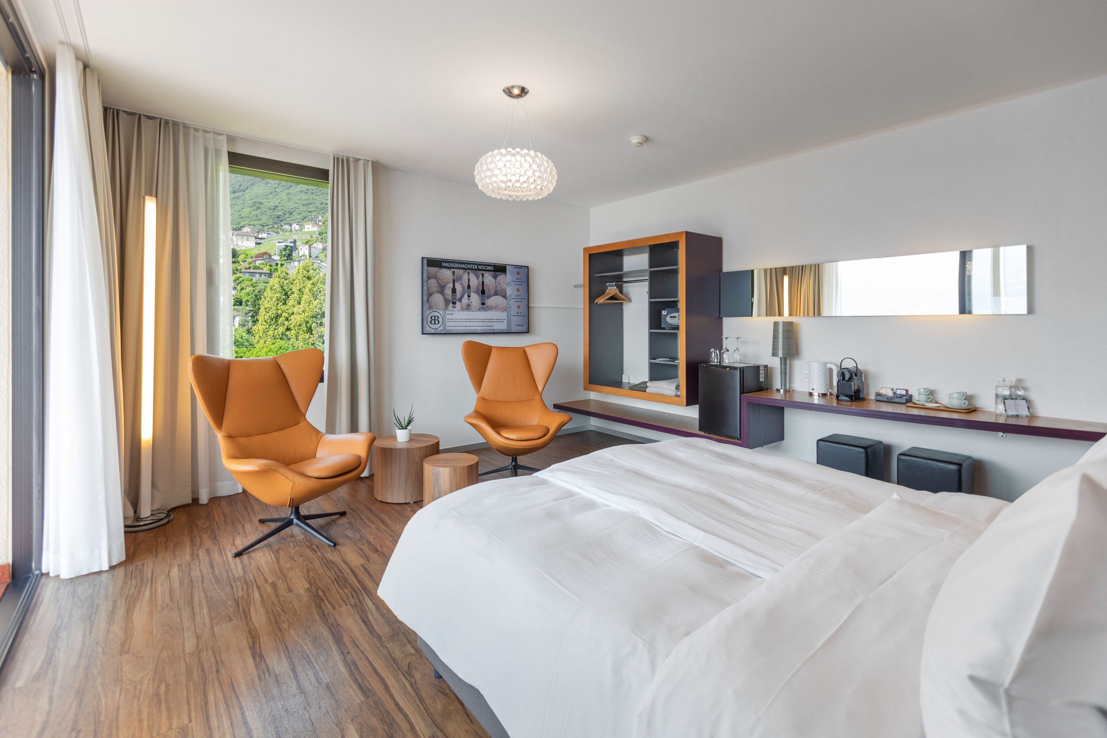Una habitación de hotel moderna con una cama grande, dos sillones, una mesita de madera, un espejo y vistas a un paisaje verde.