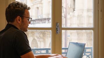 A person sitting at a wooden table with a laptop, in front of a window with a view of a building with arches.