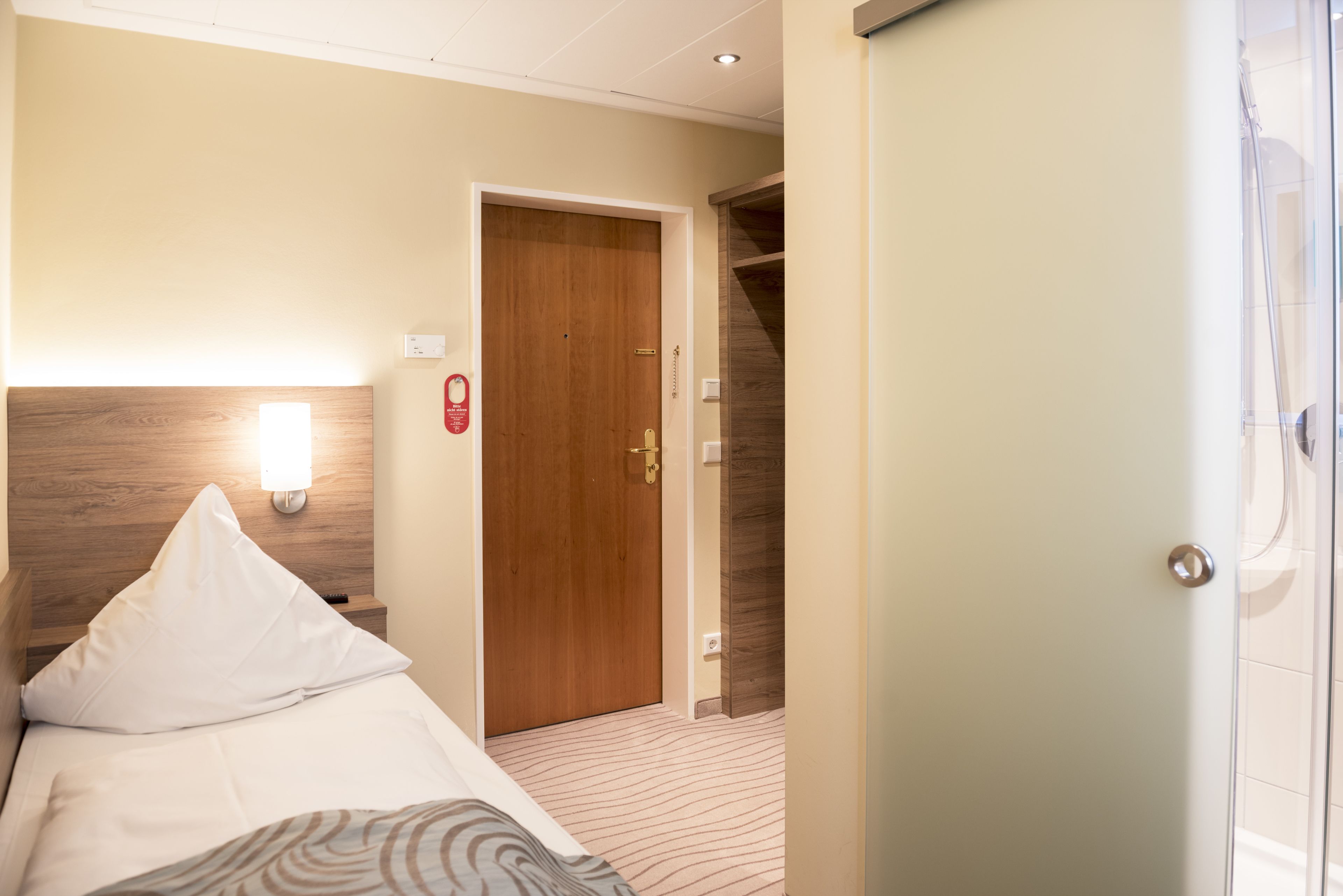 A hotel room with a bed, a nightstand, a wall-mounted light, a wooden door, and a shower area.