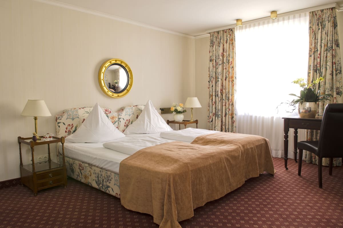 Habitación de hotel con una cama grande, dos mesitas de noche con lámparas, un espejo, un escritorio con una silla y una ventana con cortinas.
