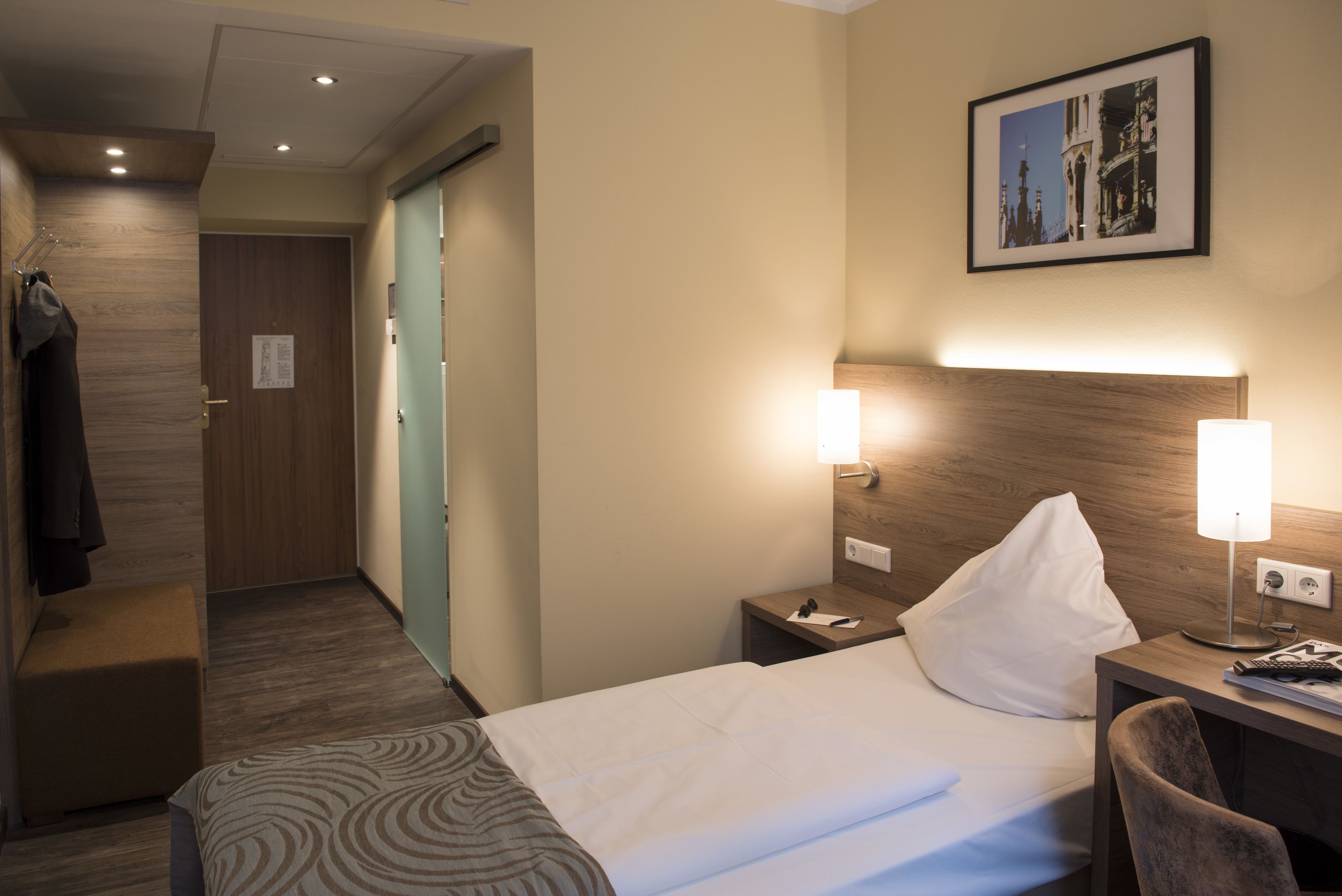 Ein Hotelzimmer mit Bett, Nachttischen, einem Sessel, Schreibtisch, Lampe, einem gerahmten Bild an der Wand und einer Tür zu einem anderen Raum.