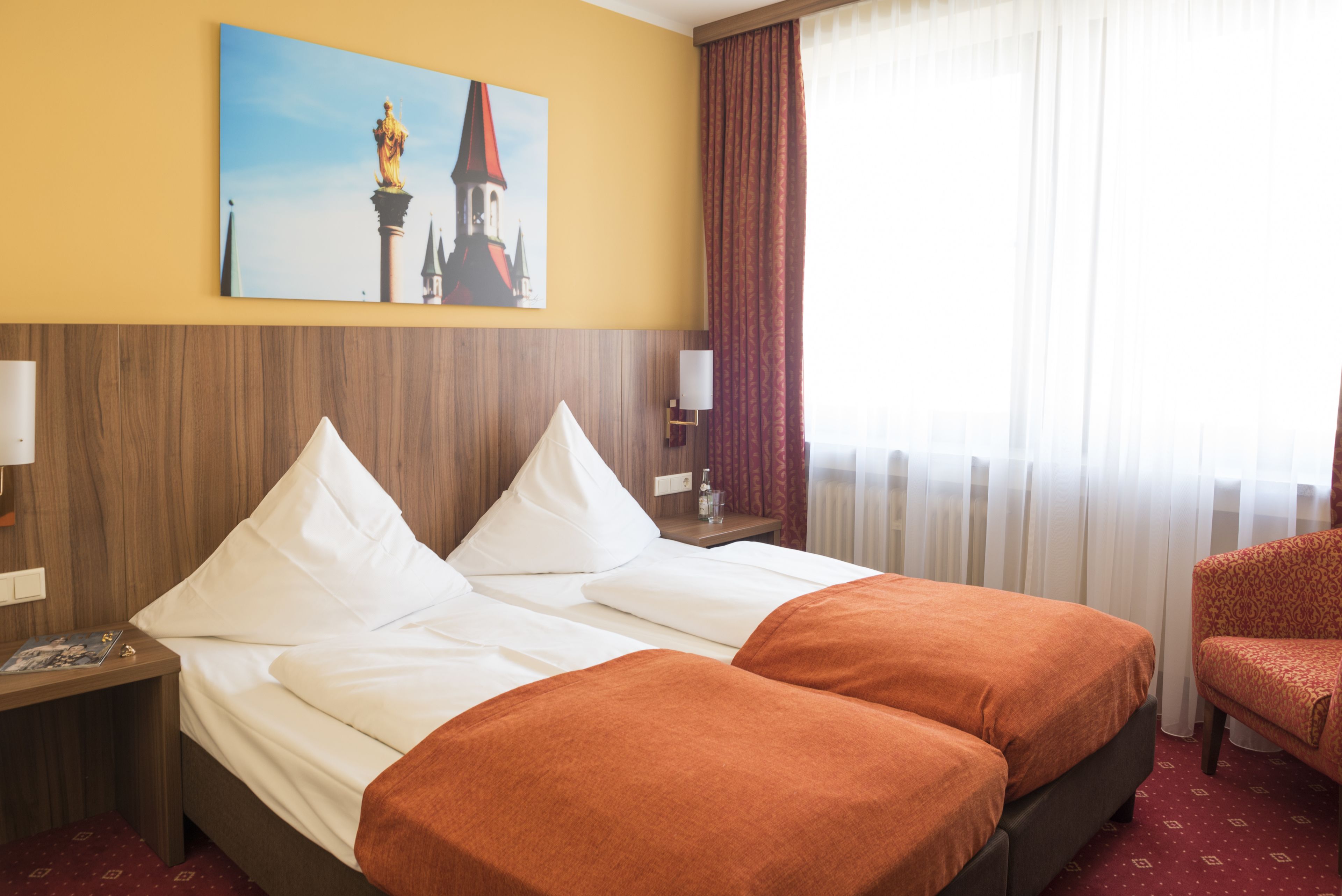 Camera d'albergo con due letti, una testiera di legno, un quadro raffigurante una torre, una sedia rossa e una finestra con tende.