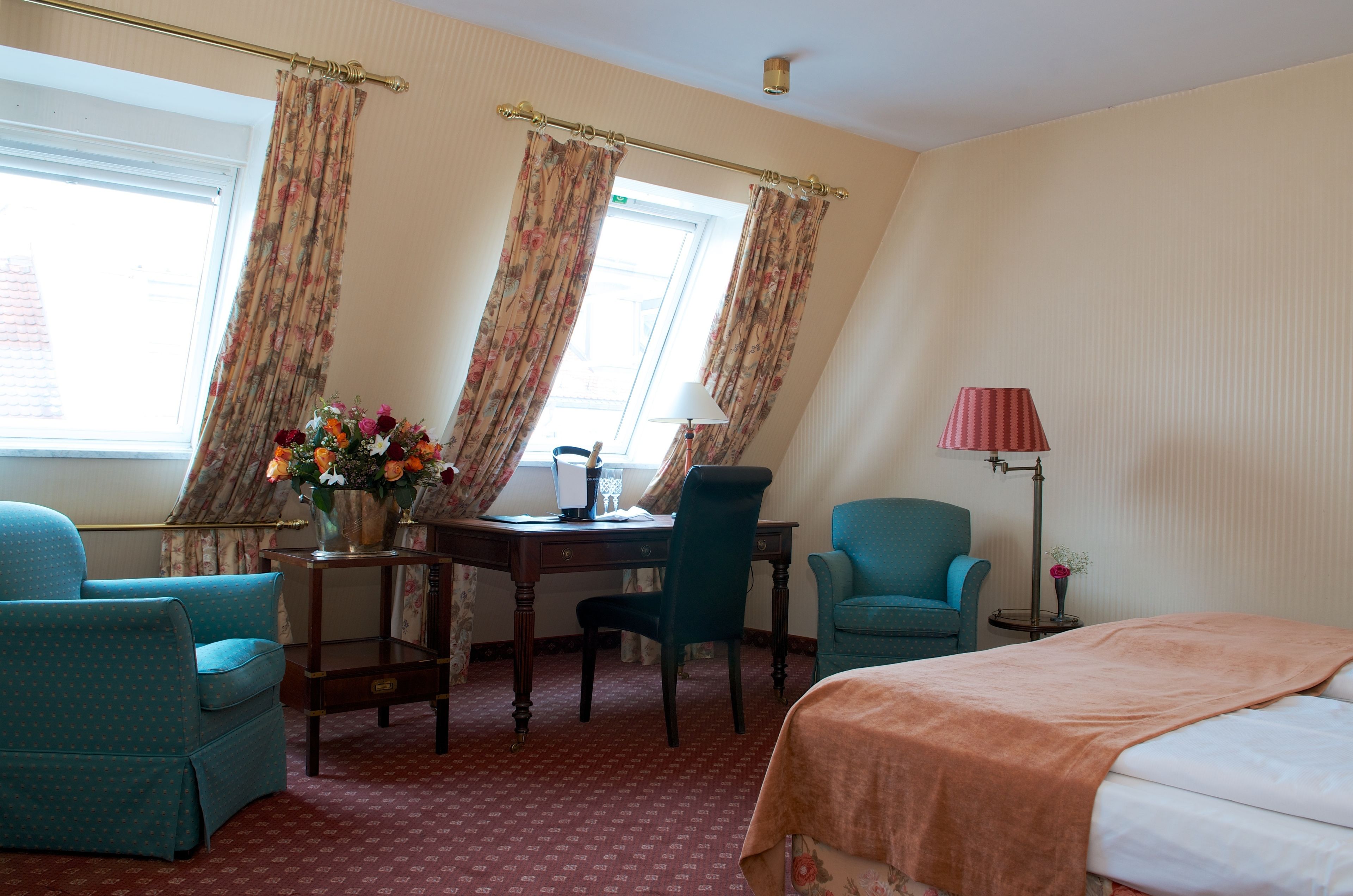 A hotel room with a bed, two armchairs, a desk with a lamp, a vase with flowers, and curtains.