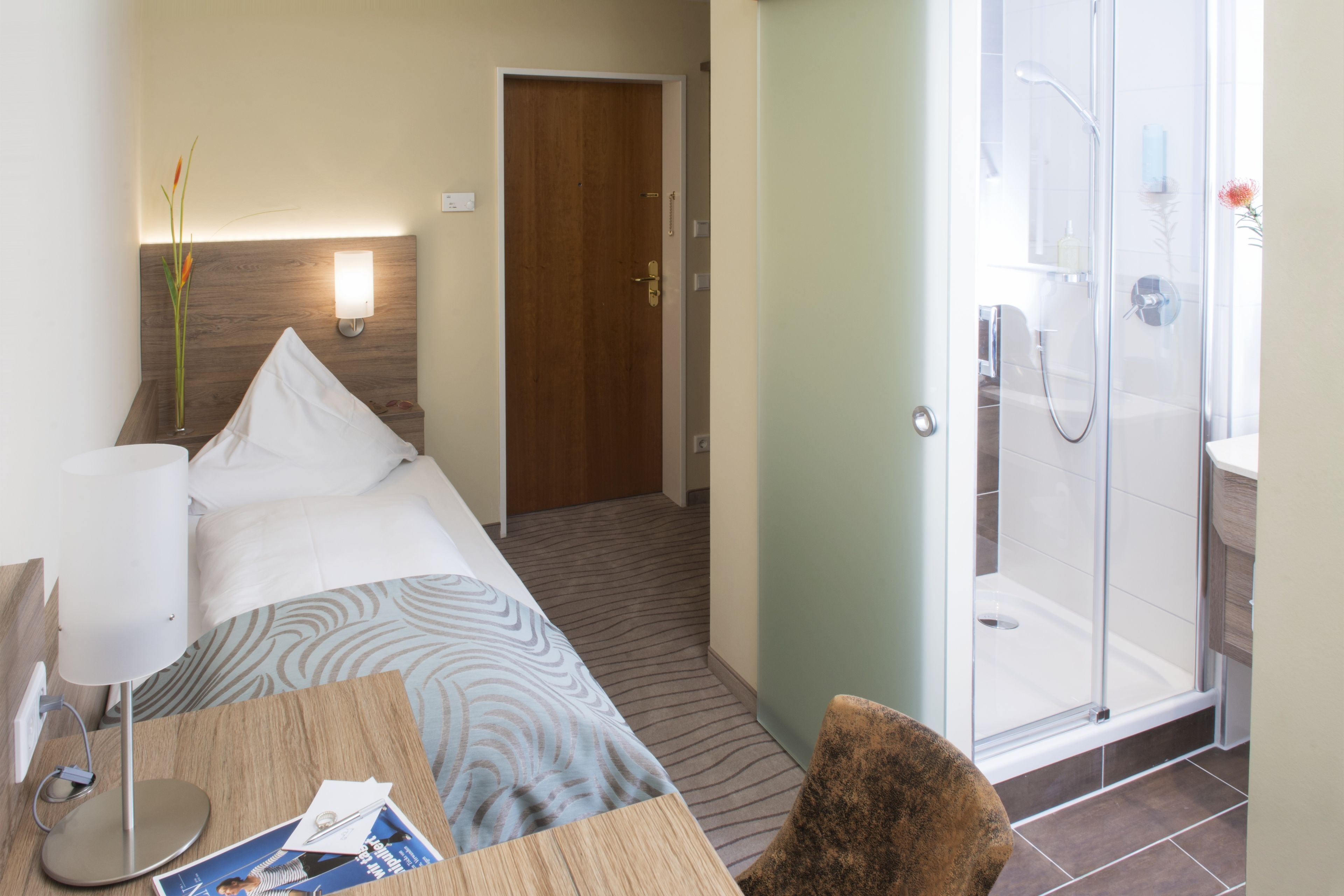 Habitación de hotel con cama, mesita de noche, lámpara y zona de ducha.